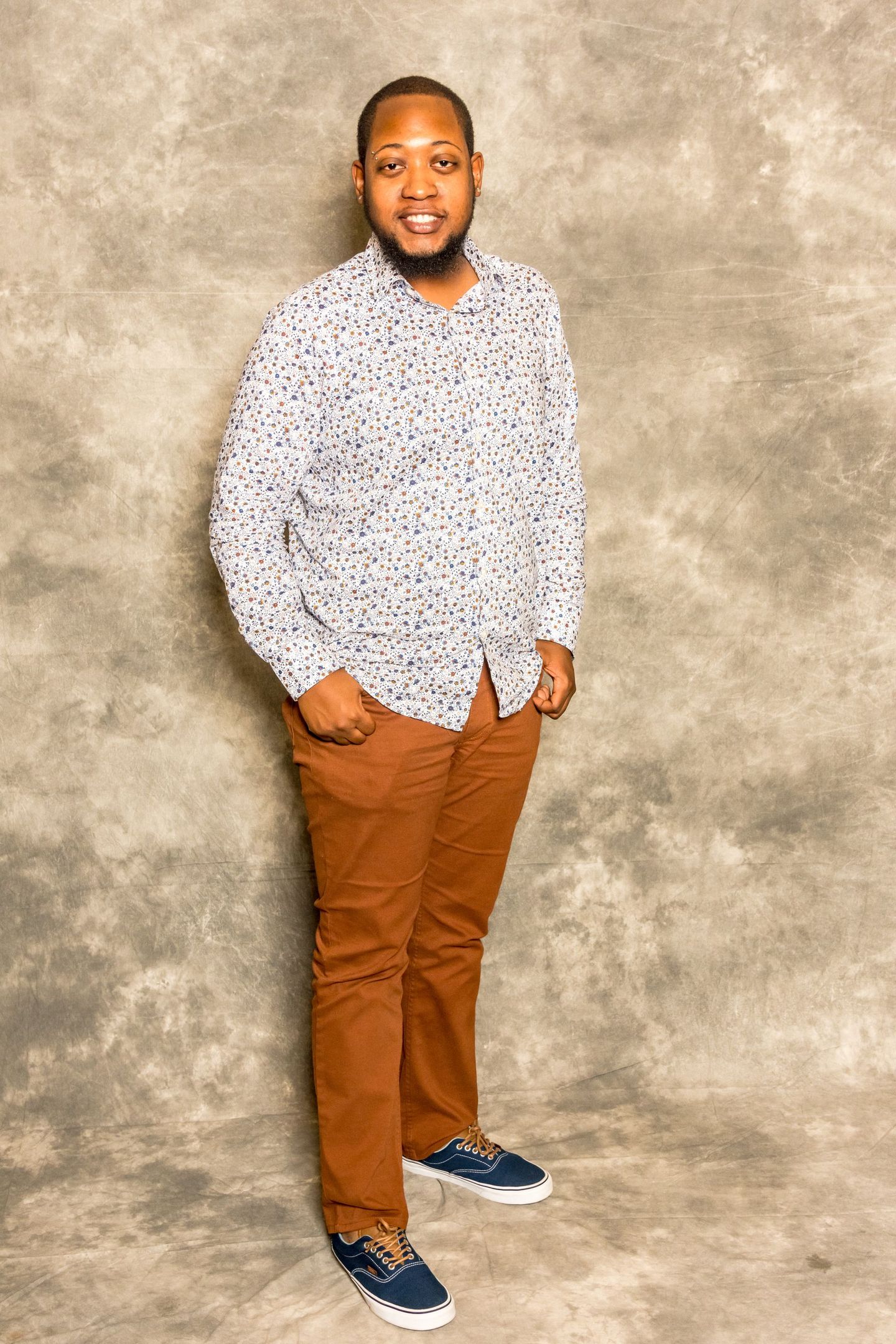 Person standing against a gray backdrop, wearing a patterned shirt, brown pants, and blue sneakers.