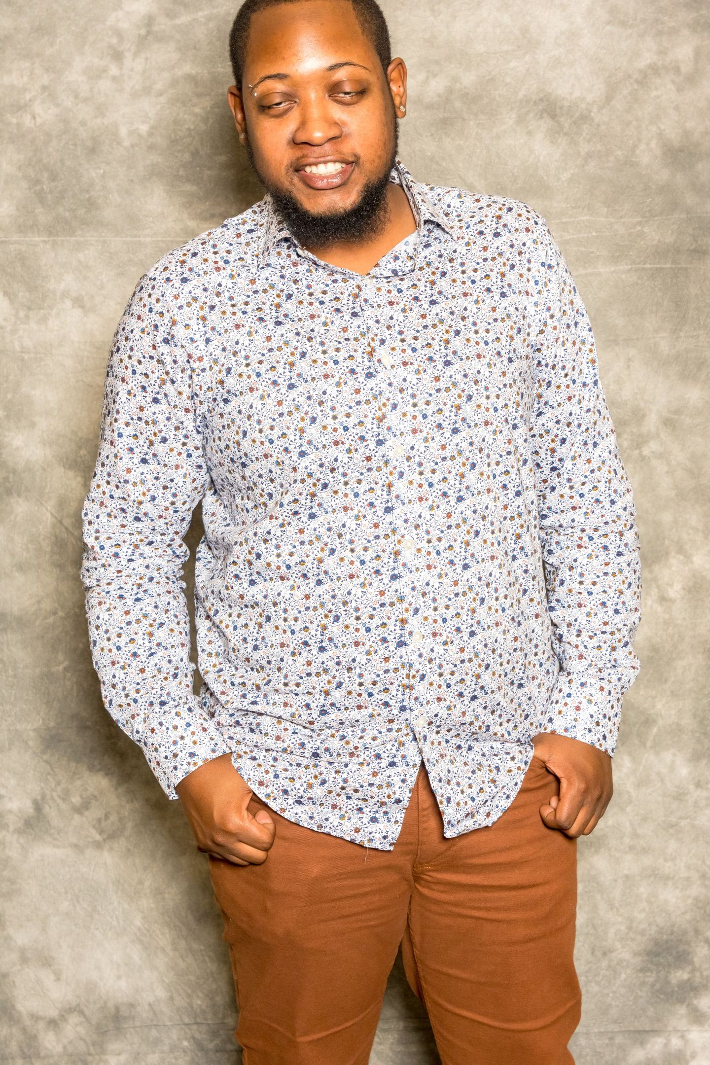 Man standing against a gray backdrop, wearing a white patterned shirt and brown pants.