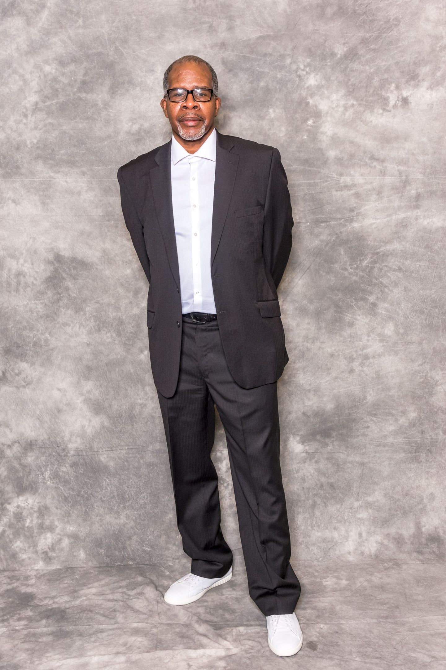 Man in dark suit and white shirt standing against a gray studio backdrop, wearing white sneakers.