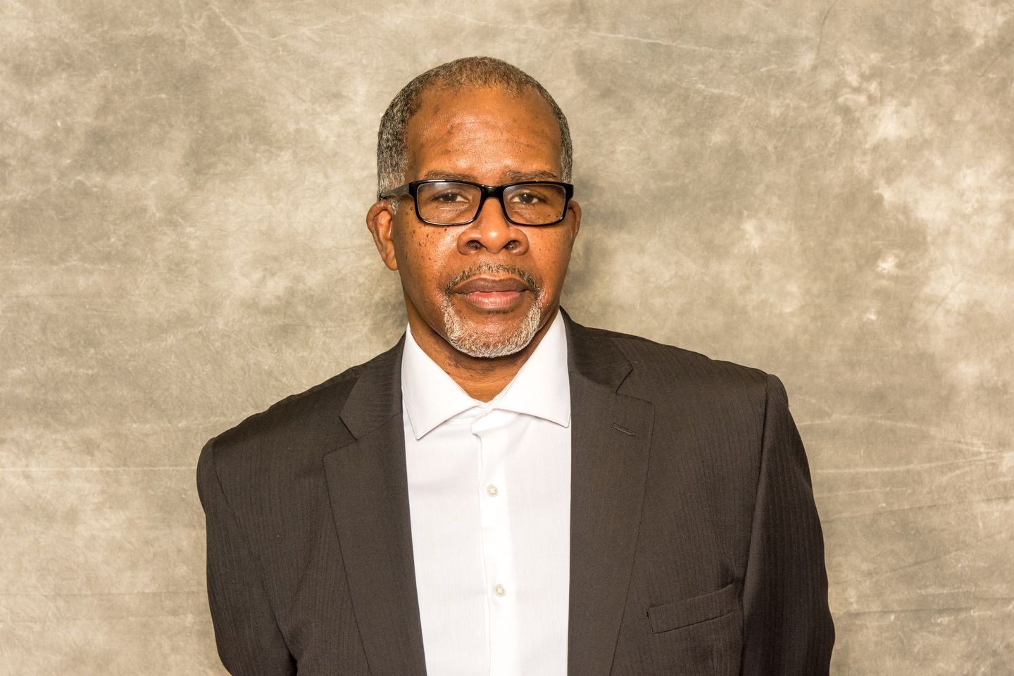 Man in a dark suit and glasses standing against a beige backdrop.
