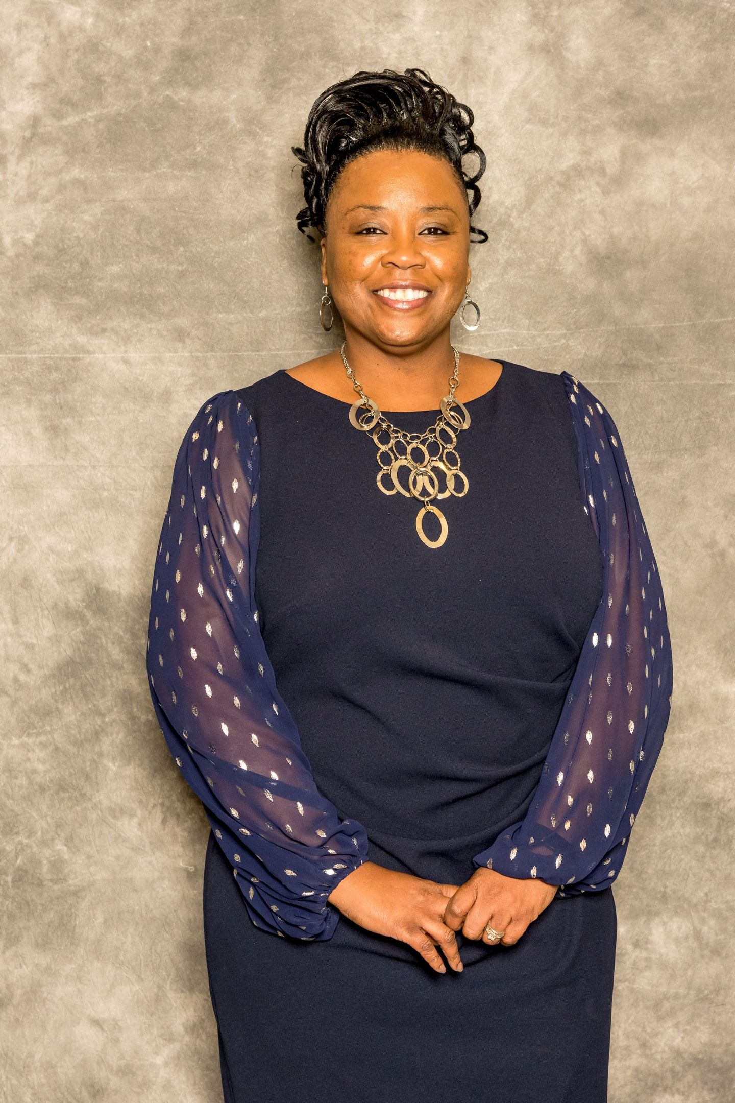 Portrait of a smiling woman in a navy dress with sheer sleeves against a textured gray backdrop