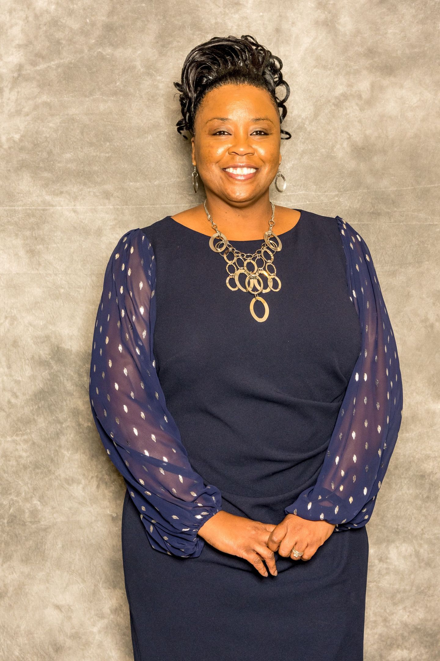 Person standing against a gray backdrop, wearing a navy dress with sheer sleeves and a gold necklace, smiling.