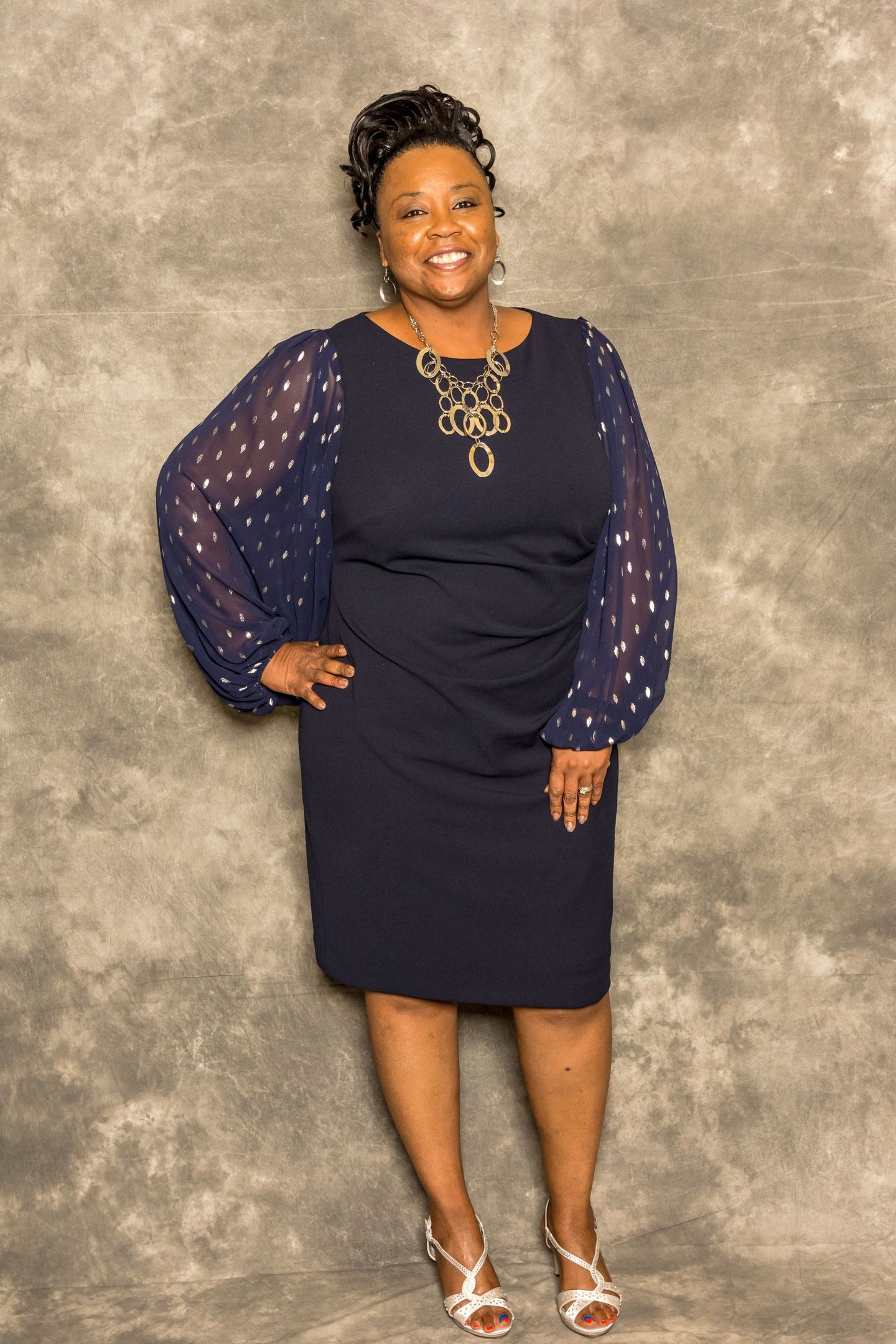 Woman in navy dress and sheer sleeves posing against a mottled gray backdrop, wearing silver heels.