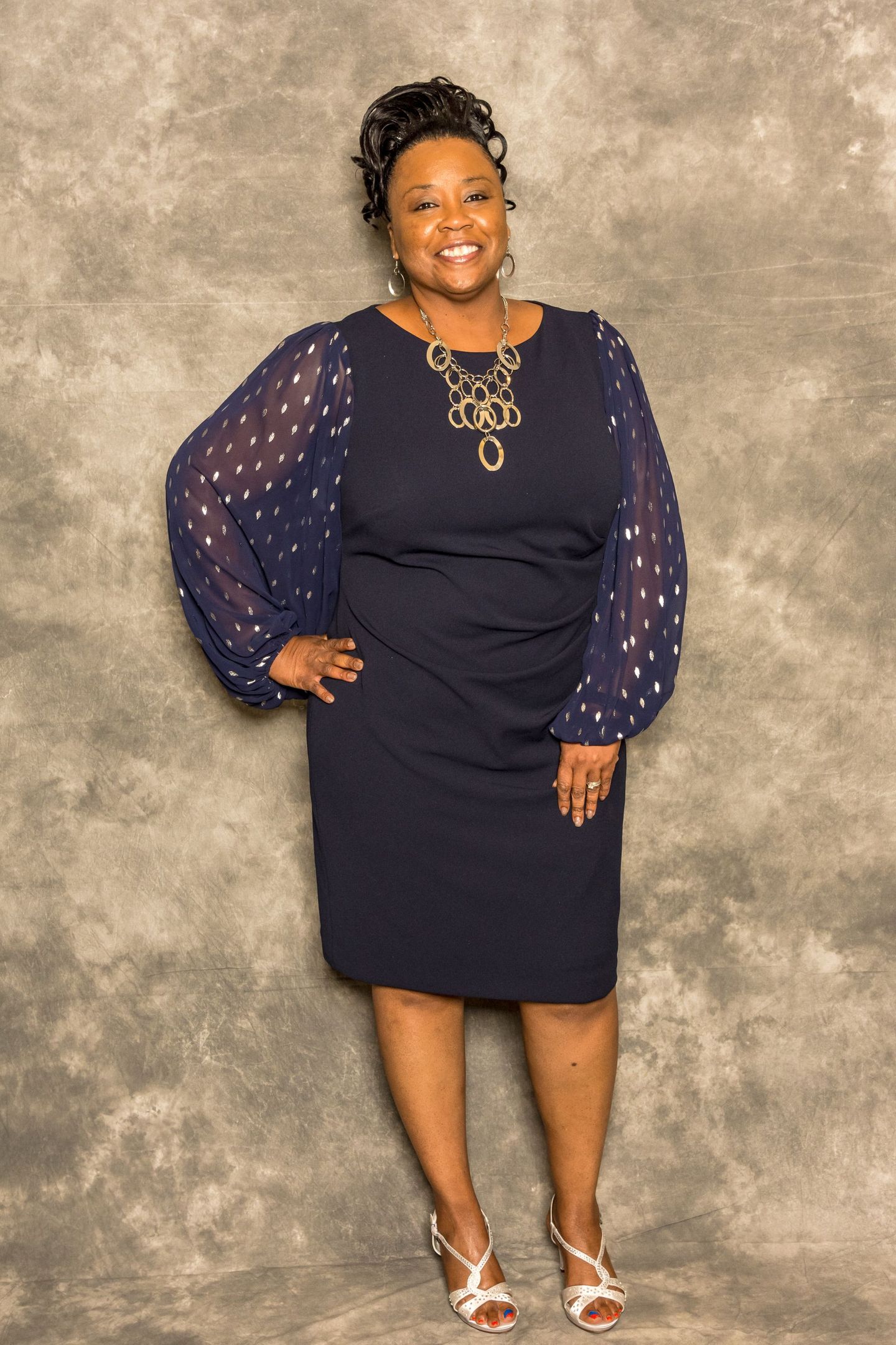 Woman in a navy dress with sheer sleeves poses against a mottled gray backdrop.