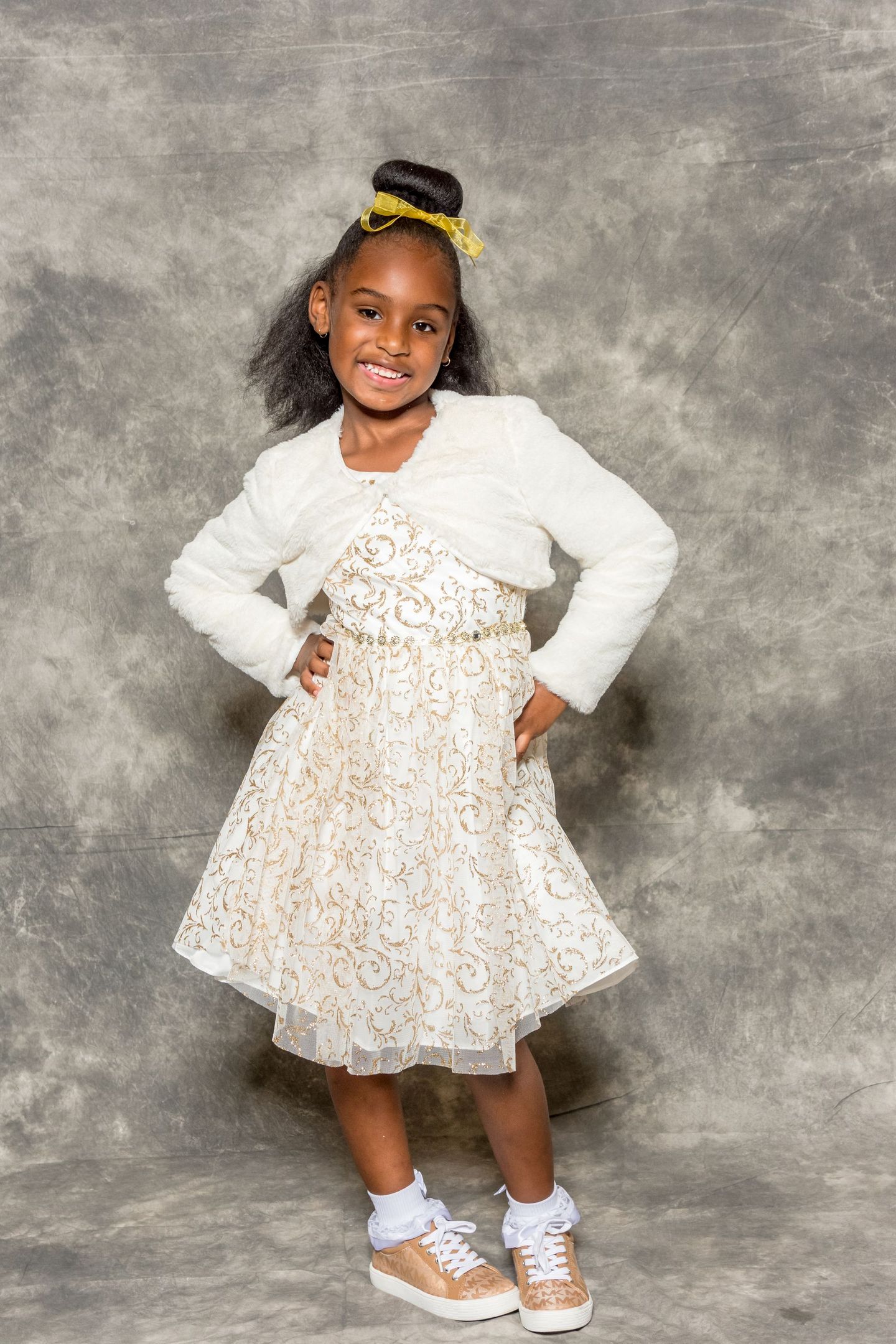 Smiling child in a white patterned dress and sandals, standing with hands on hips against a gray backdrop.