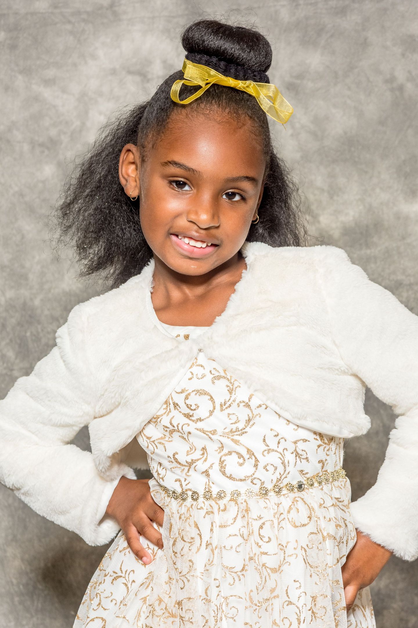 Young child in a white dress with gold accents, smiling and posing against a gray studio backdrop
