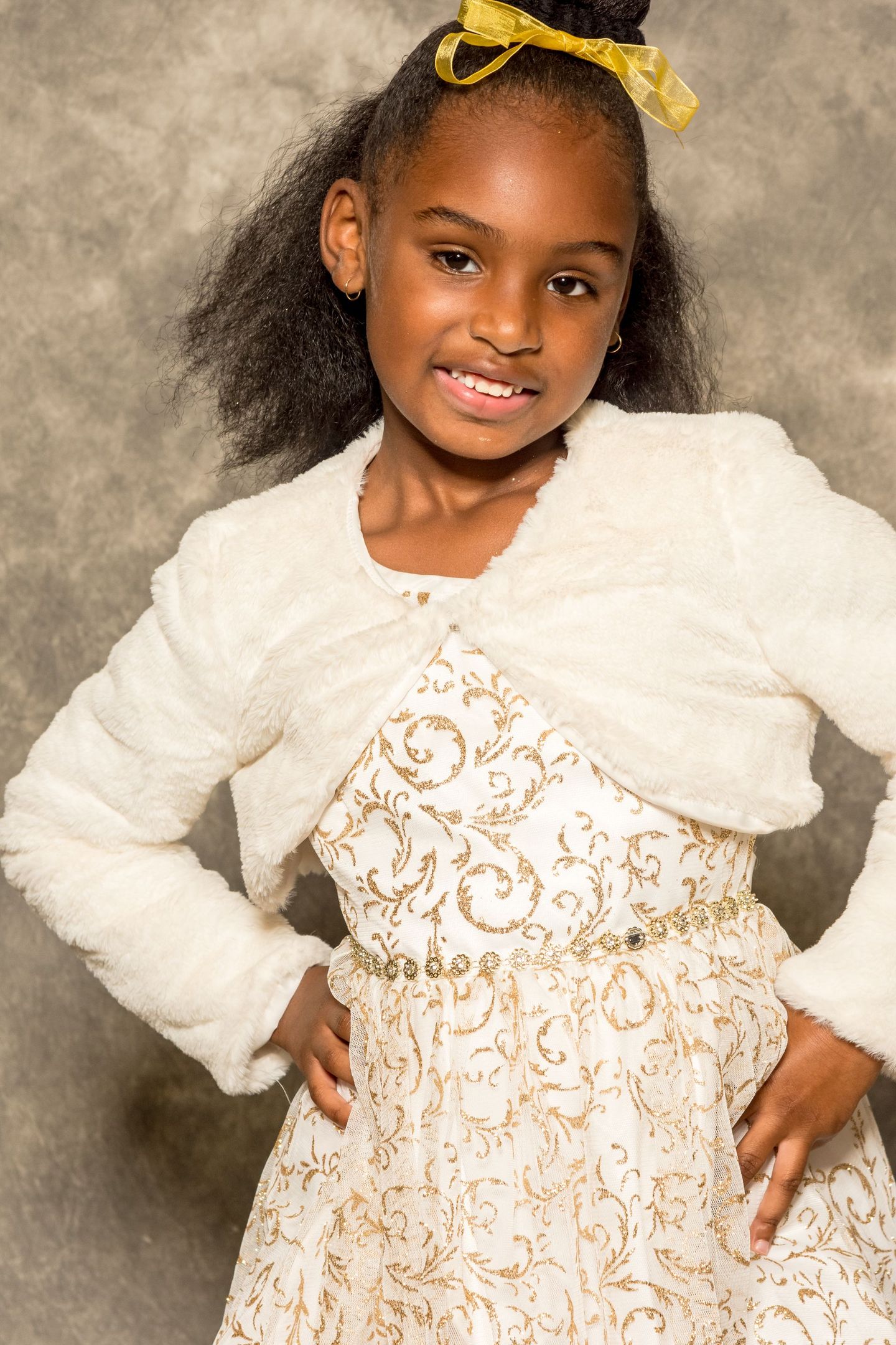 Young girl in a white dress and cardigan, smiling with hands on hips against a studio backdrop