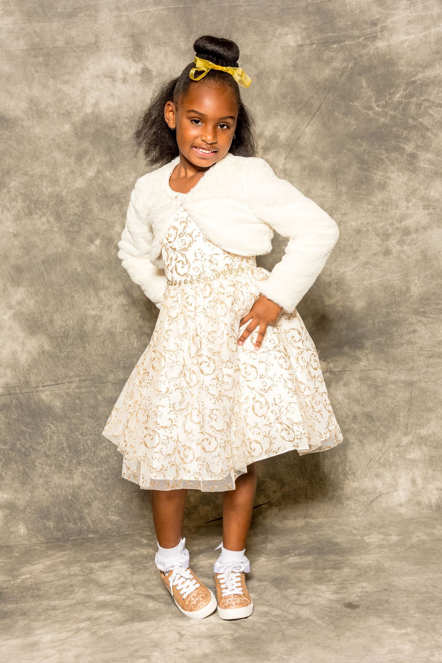 Young child in a white floral dress and cardigan posing against a gray studio backdrop.