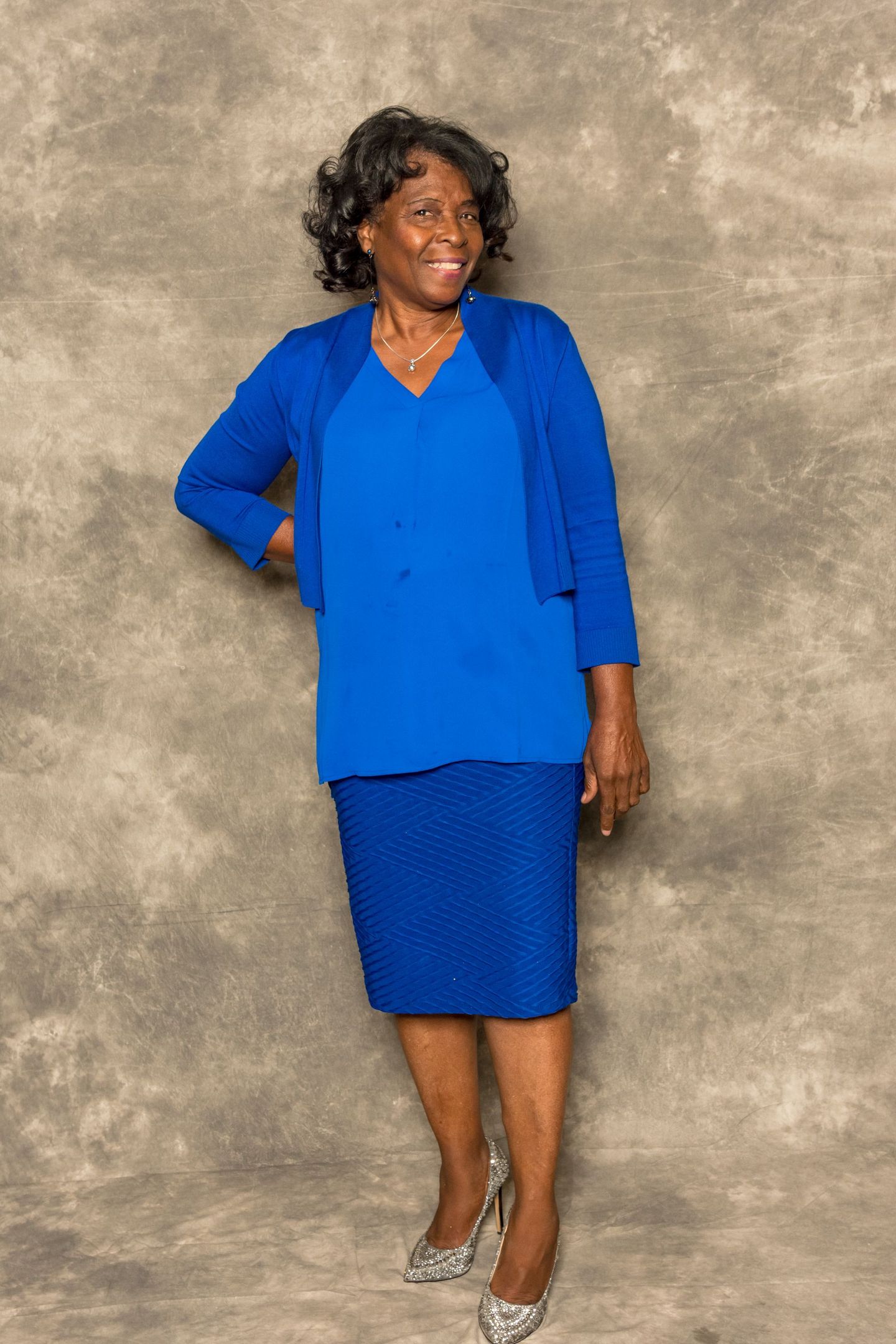 Woman standing against a mottled backdrop, wearing a bright blue blouse and skirt with silver heels.