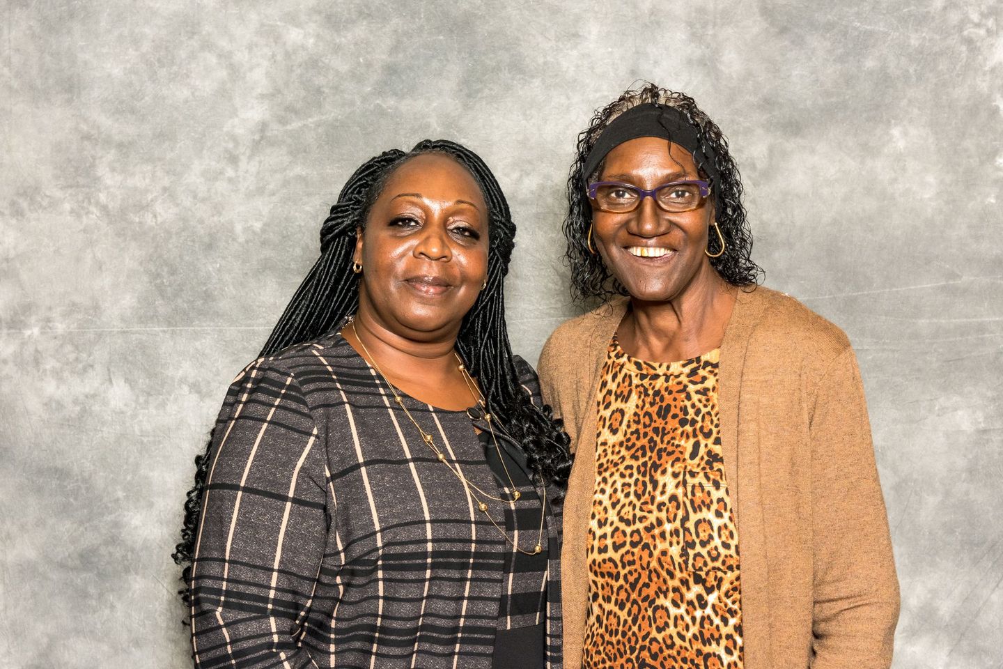 Two women standing together, smiling, against a gray textured backdrop.