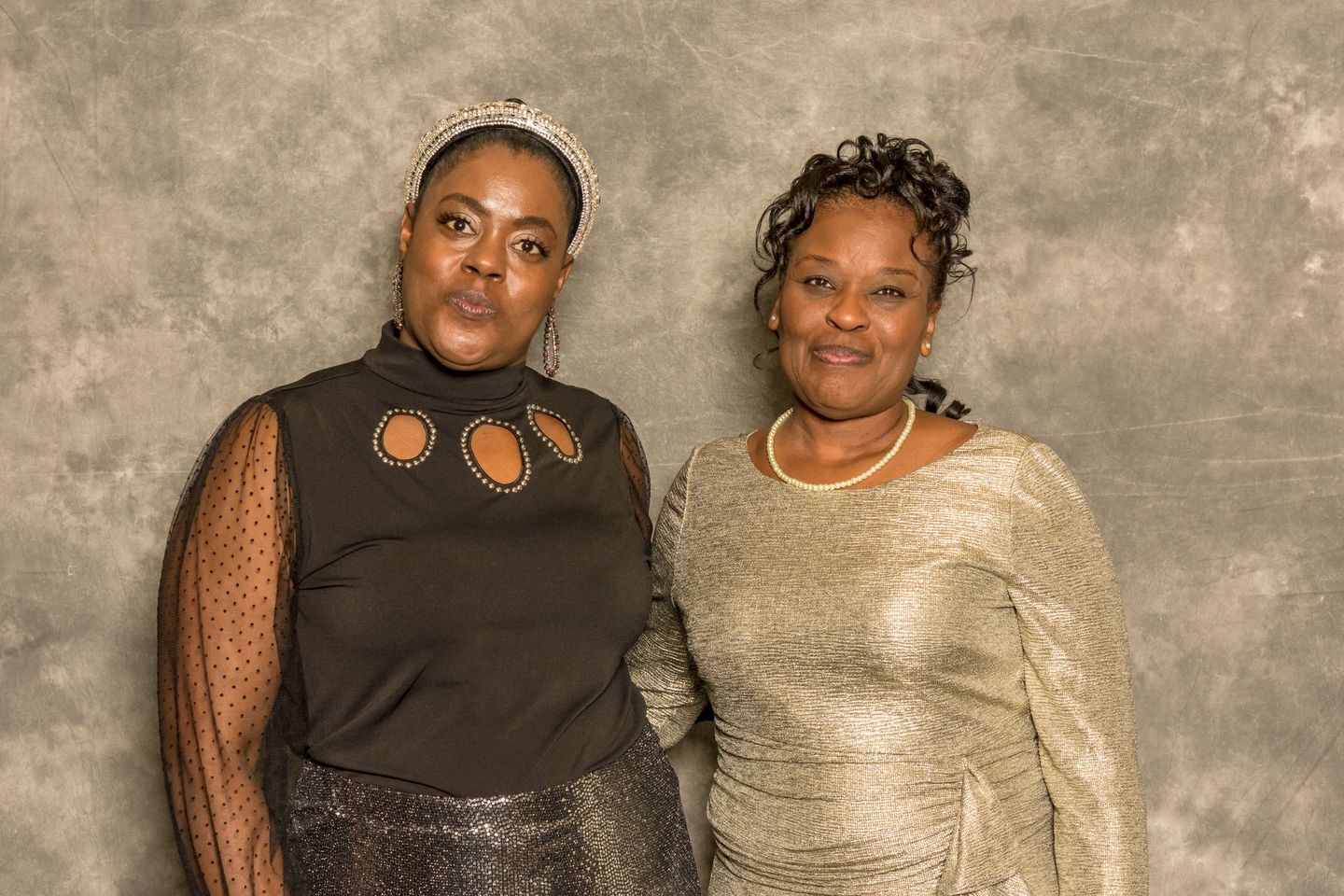 Two women posing together against a textured gray backdrop, wearing formal tops and smiling.