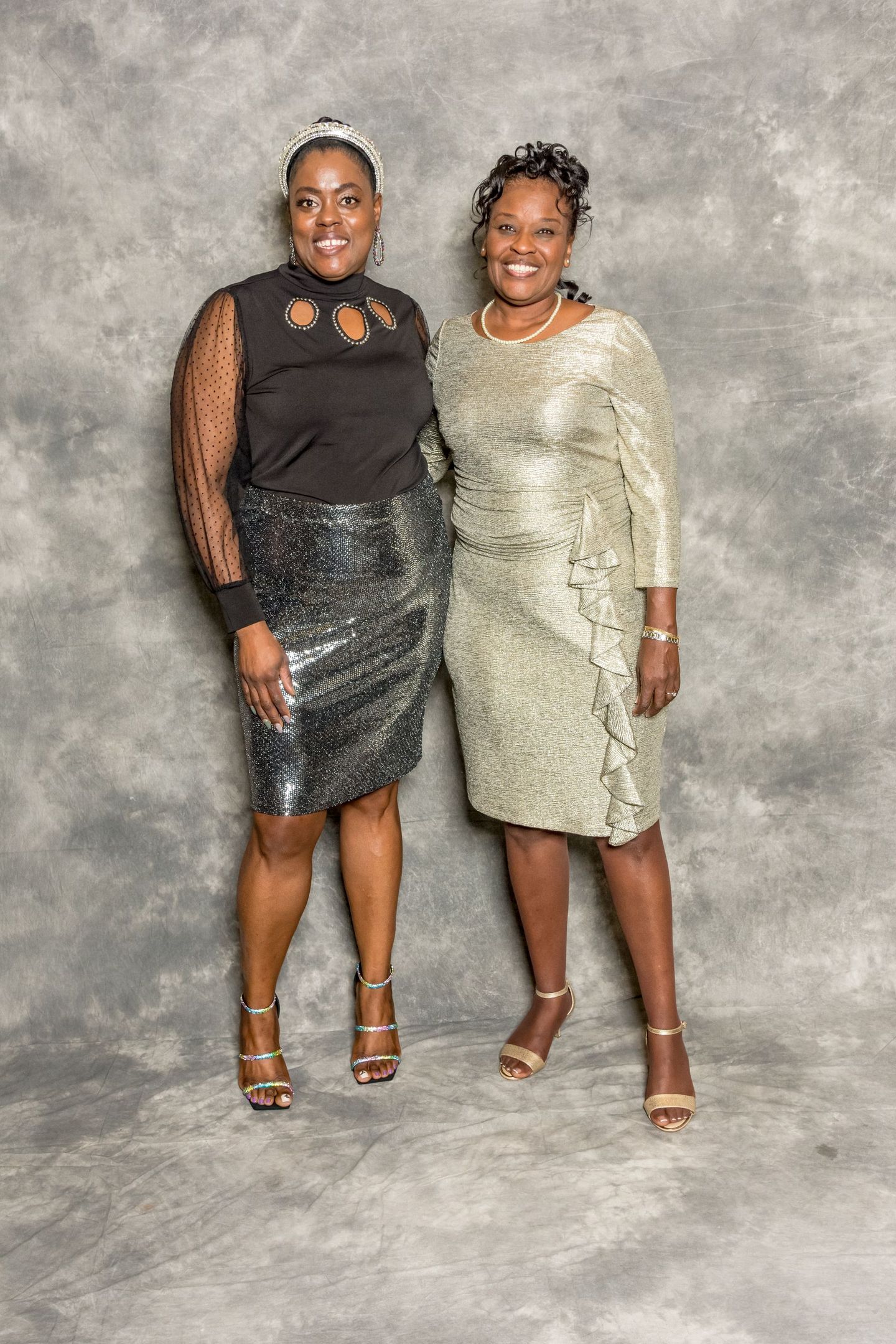 Two women posing together in elegant black and silver dresses against a gray backdrop.