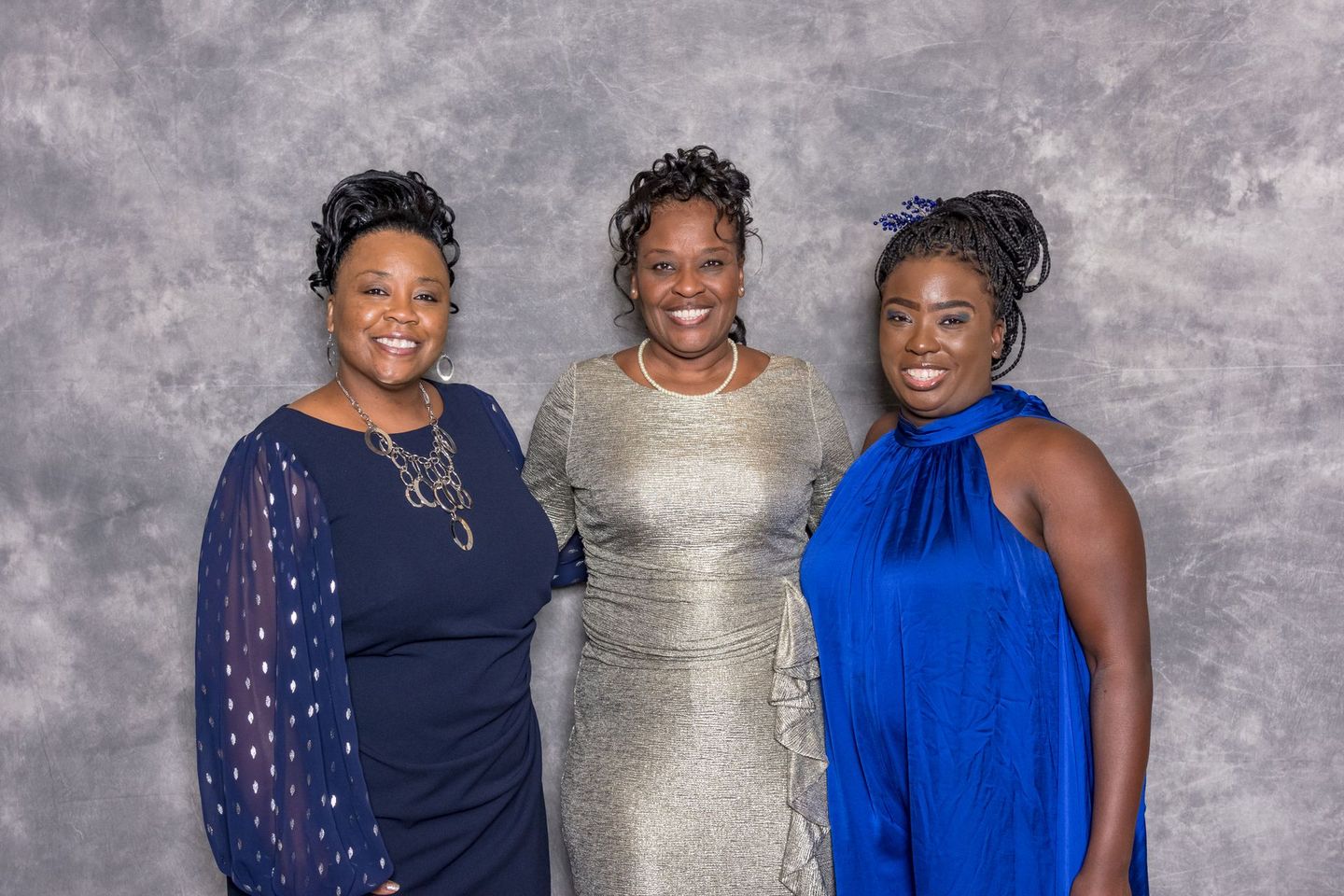 Three women pose smiling in formal dresses against a gray studio backdrop.