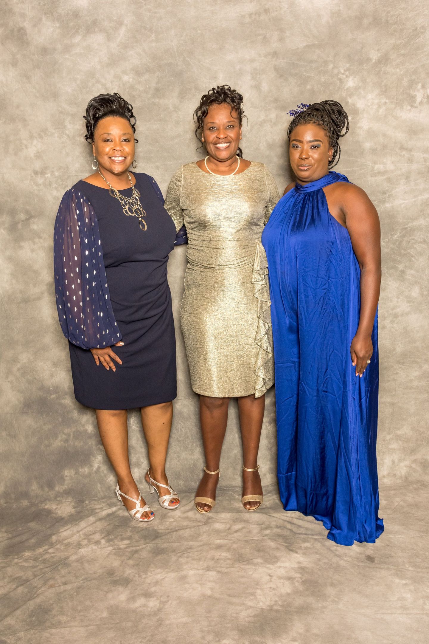 Three women standing together in formal dresses against a neutral backdrop, smiling at the camera.