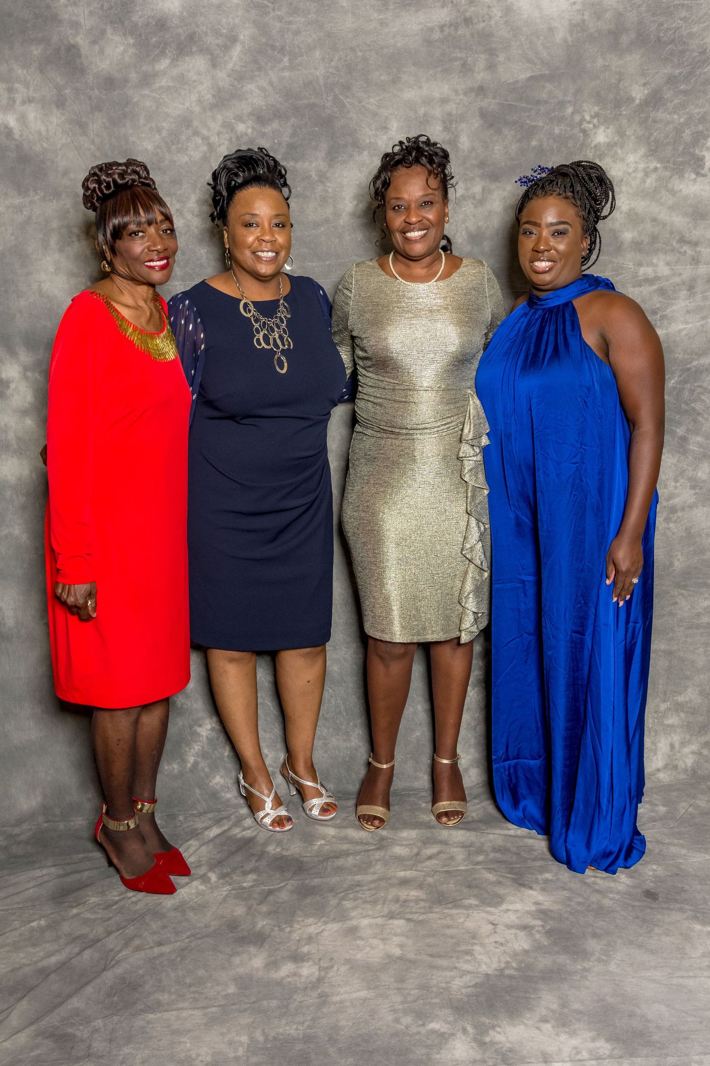 Four women in formal dresses pose together against a gray backdrop.