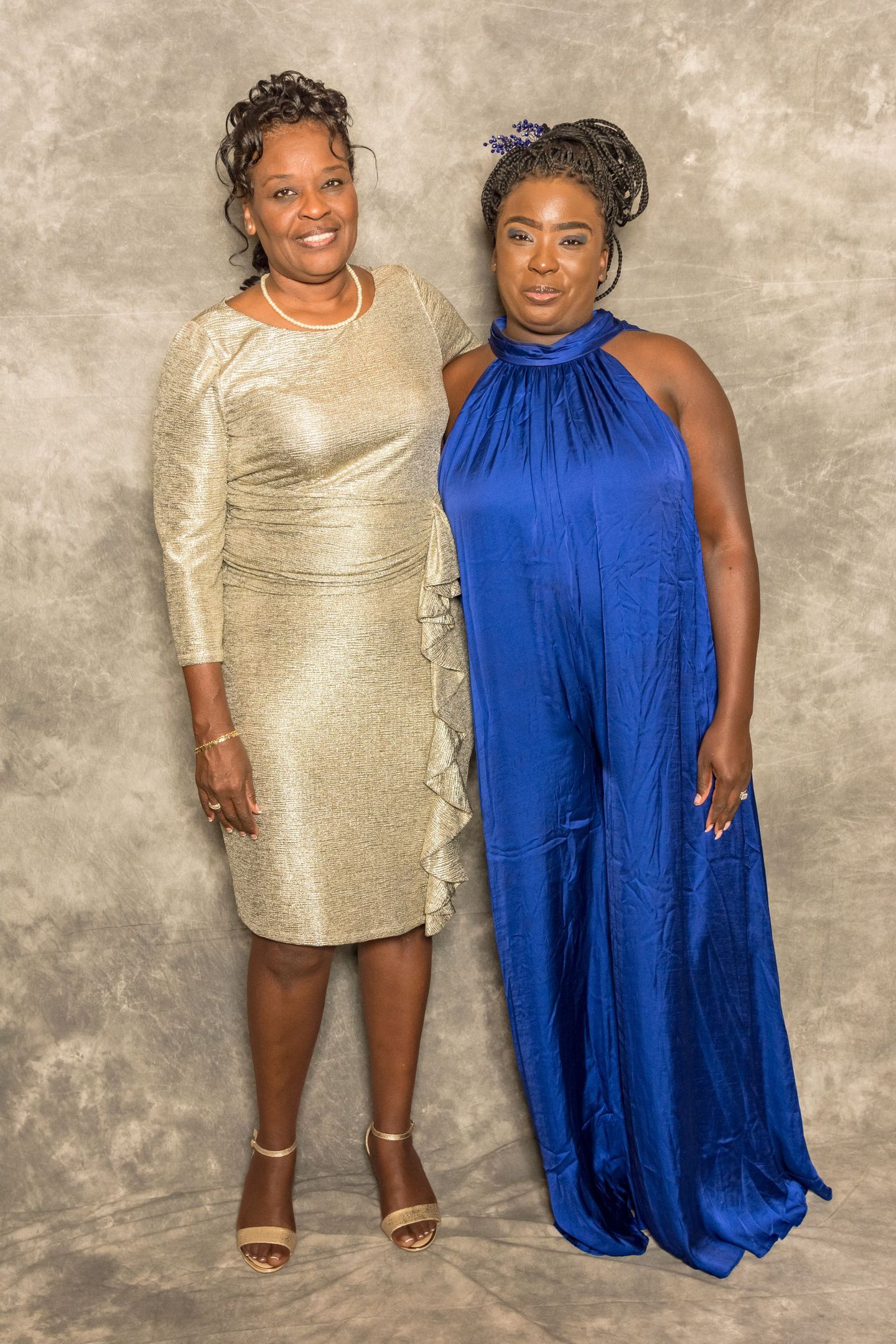 Two women posing in formal dresses, one gold and one blue, against a textured backdrop.