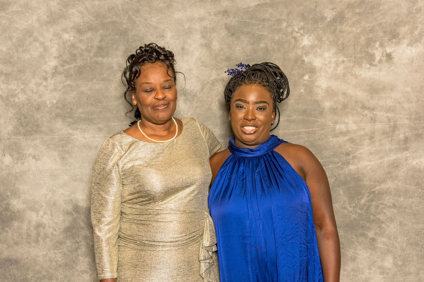 Two people posing together in front of a gray backdrop, wearing gold and blue dresses
