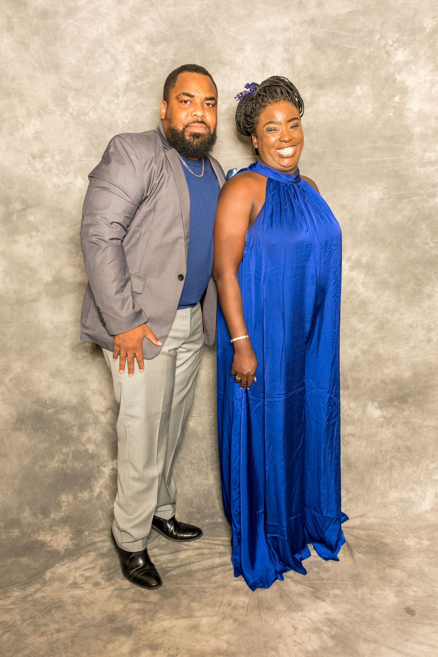 Two people posing against a textured beige backdrop; one in a gray suit, the other in a royal blue dress.