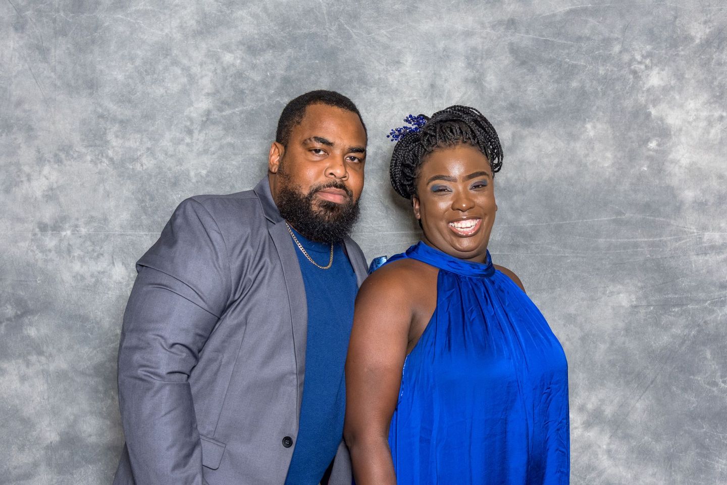 Two people posing together against a gray backdrop, one in a gray jacket and blue shirt, the other in a blue dress.