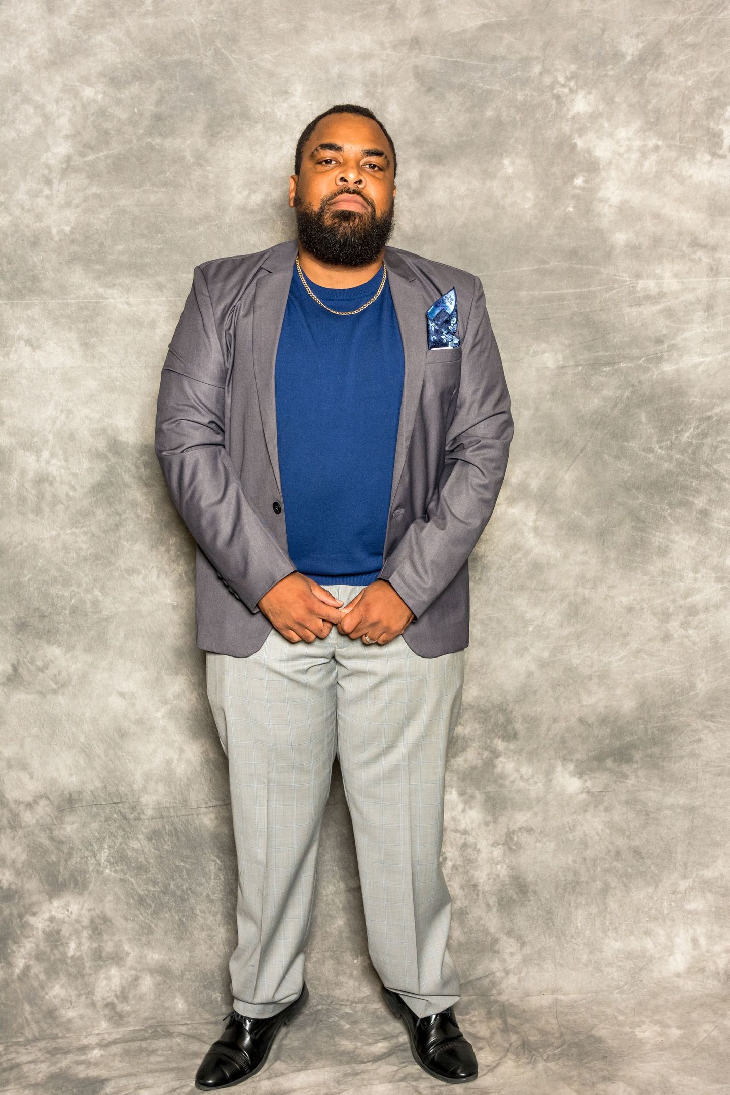 Man in gray blazer and light pants standing against a gray backdrop, hands clasped in front.