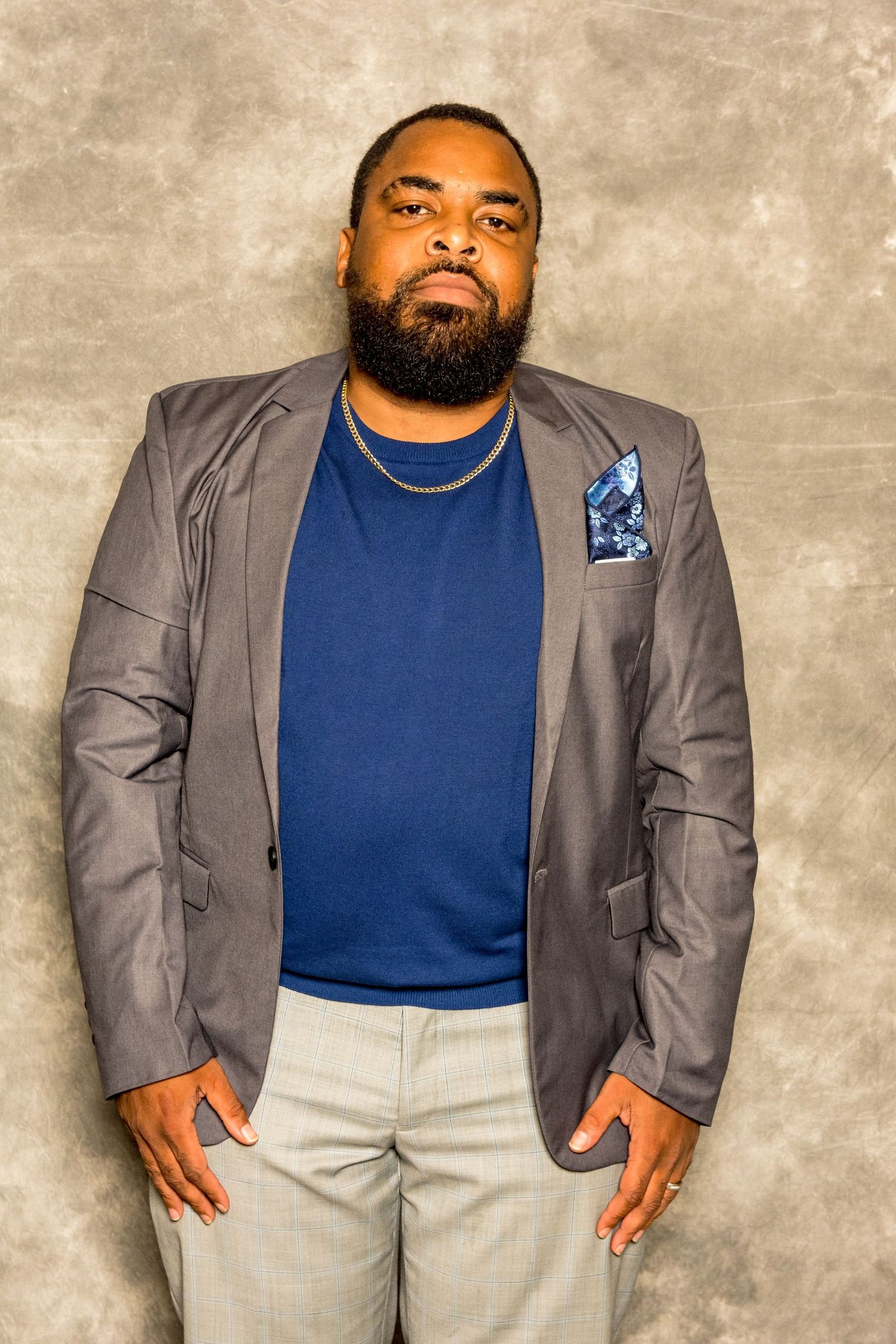 Man standing against a gray backdrop, wearing a navy shirt, gray blazer, and light pants.