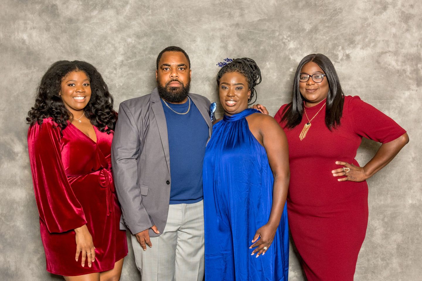 Four people pose together against a gray backdrop, wearing formal outfits in red, gray, blue, and red.
