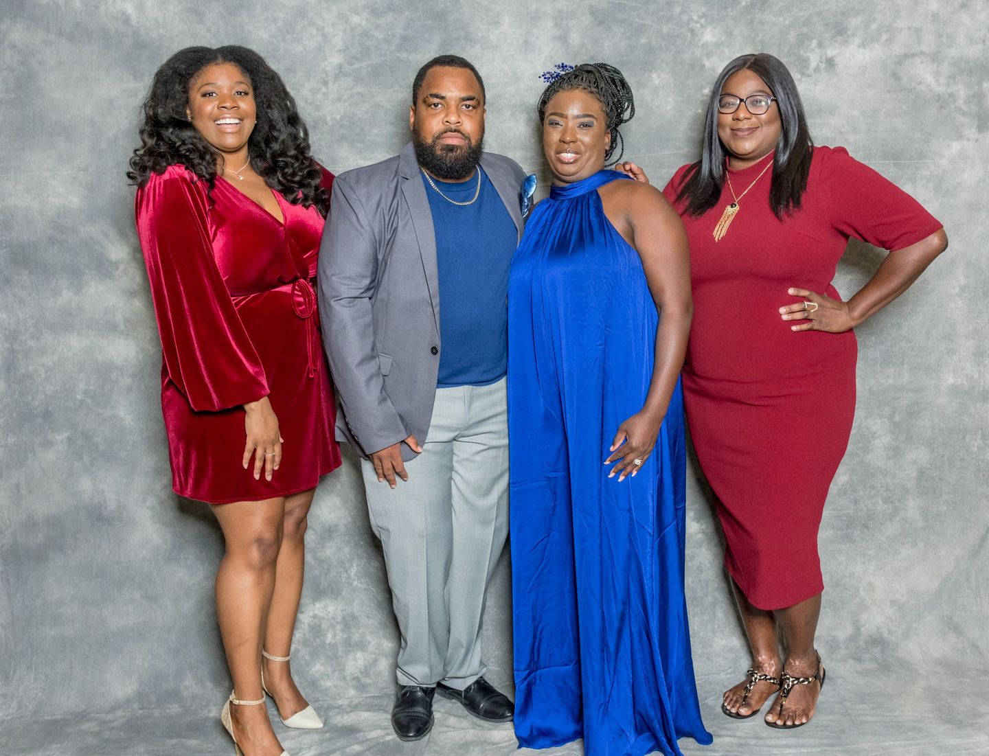 Four people posing together against a gray backdrop, dressed in red, gray, and blue formal outfits.