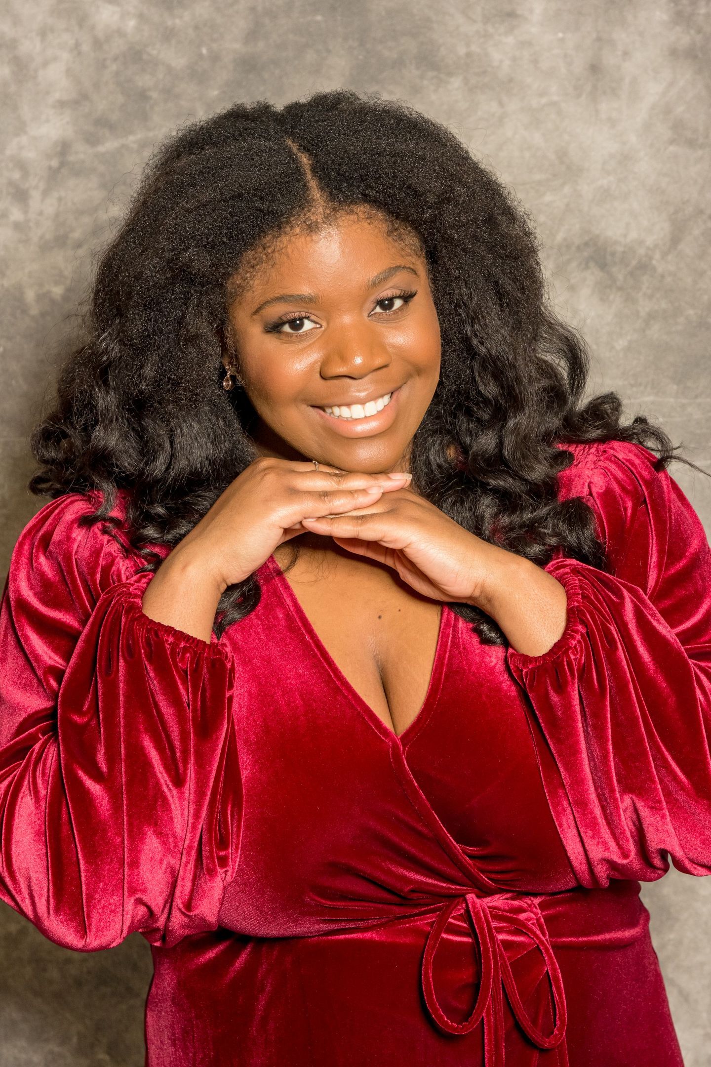 Smiling person in a red velvet top, resting chin on hands against a neutral backdrop
