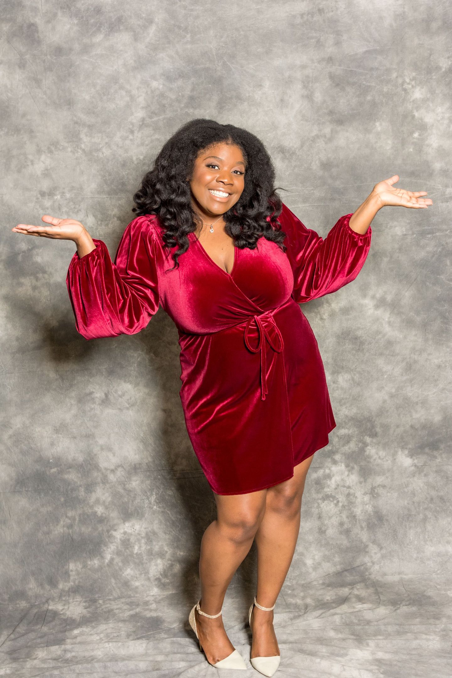 Woman in a red velvet dress posing with raised hands against a gray backdrop.