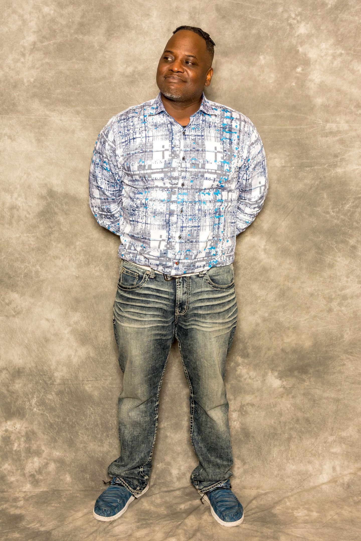 Man standing against a mottled beige backdrop, wearing a white patterned shirt, jeans, and blue sneakers.