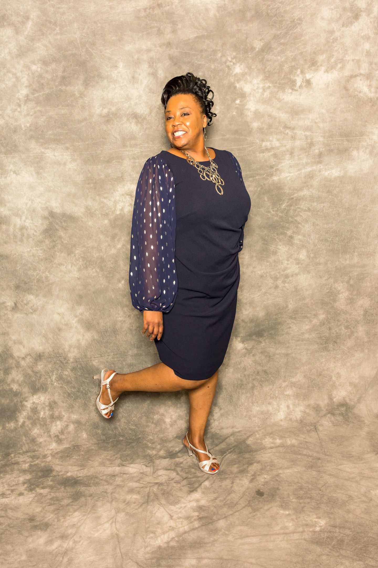 Woman in a navy dress and heeled sandals posing against a textured studio backdrop.