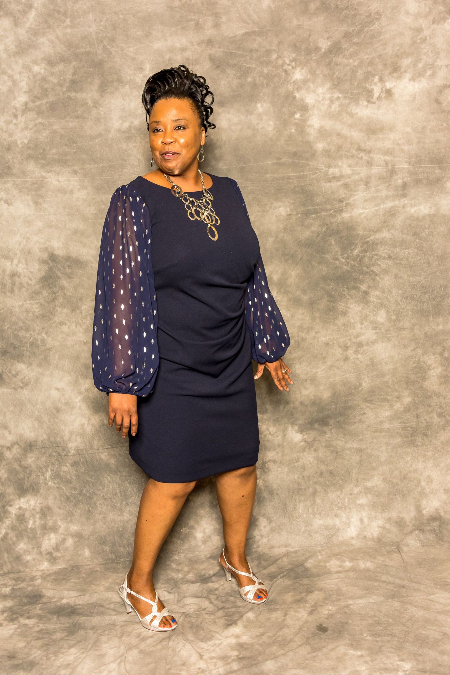 Woman in a navy dress with sheer sleeves posing against a mottled gray backdrop.