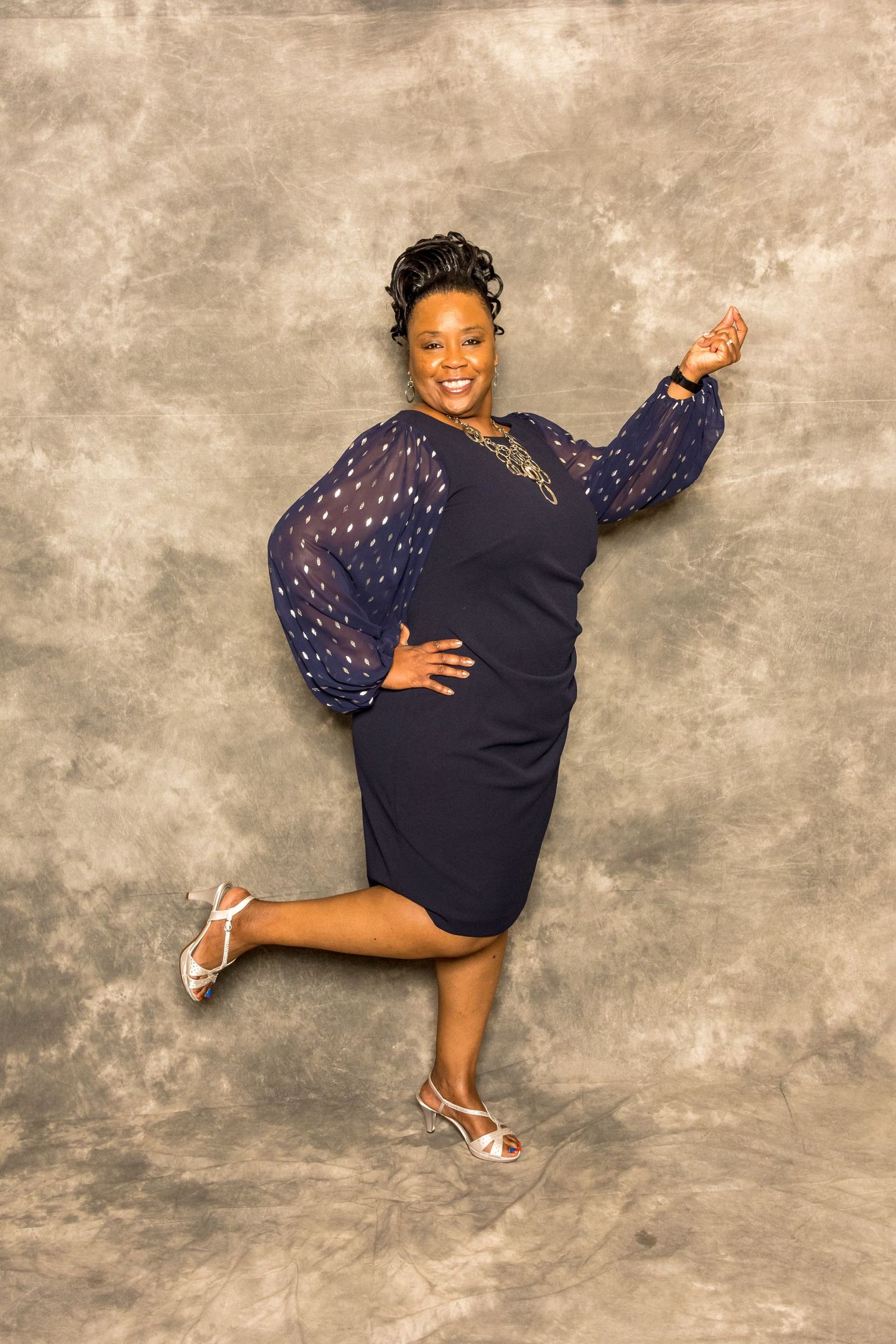 Woman in a navy dress and heels posing playfully against a textured beige backdrop.