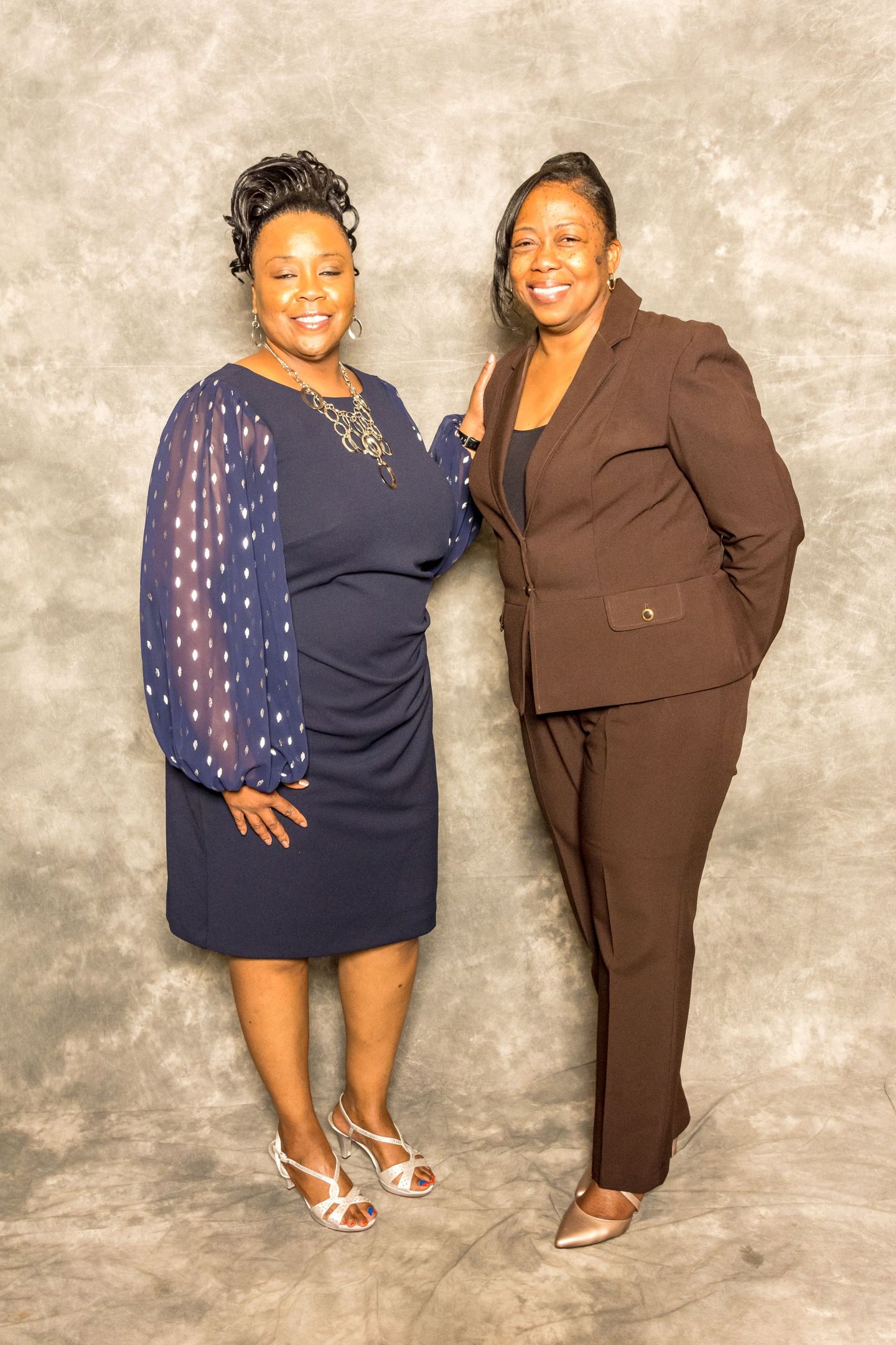Two women posing together against a gray backdrop, one in a navy dress and one in a brown suit.
