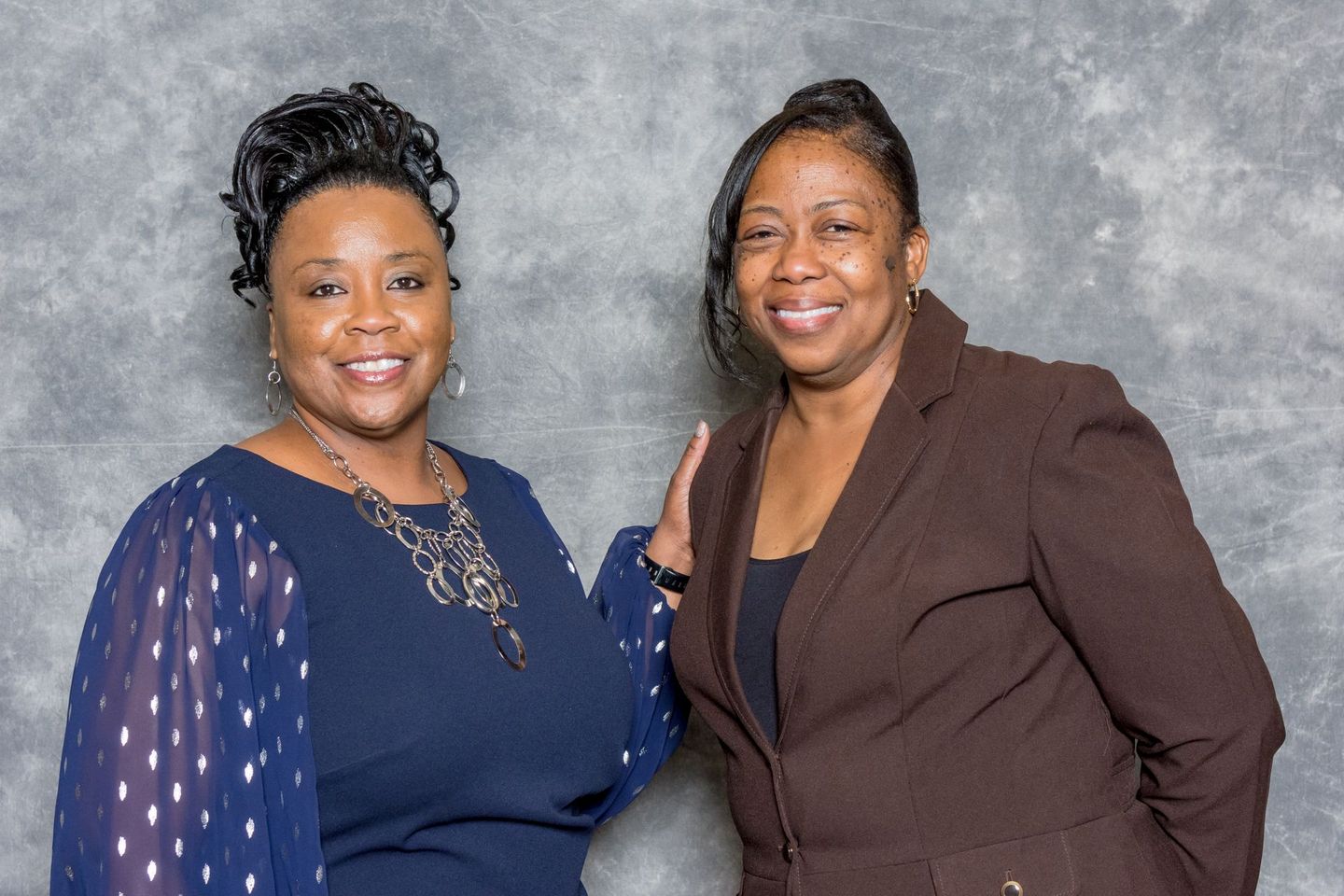 Two women smiling and posing together against a gray backdrop, wearing navy and brown outfits.