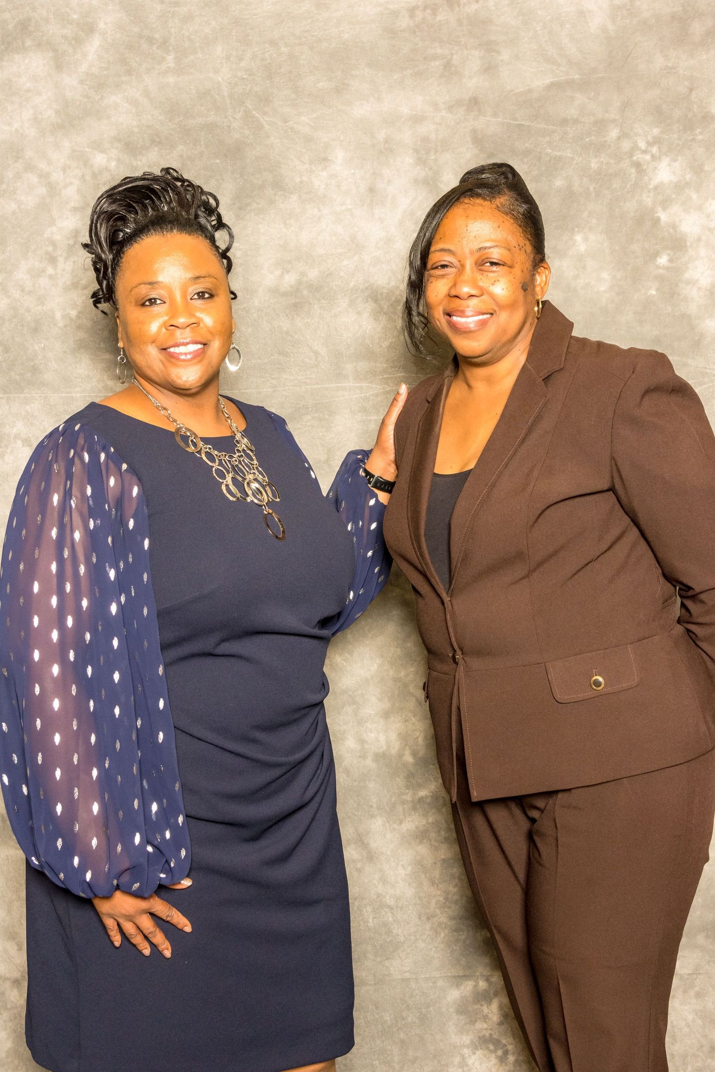Two women posing and smiling in dark outfits against a gray studio backdrop