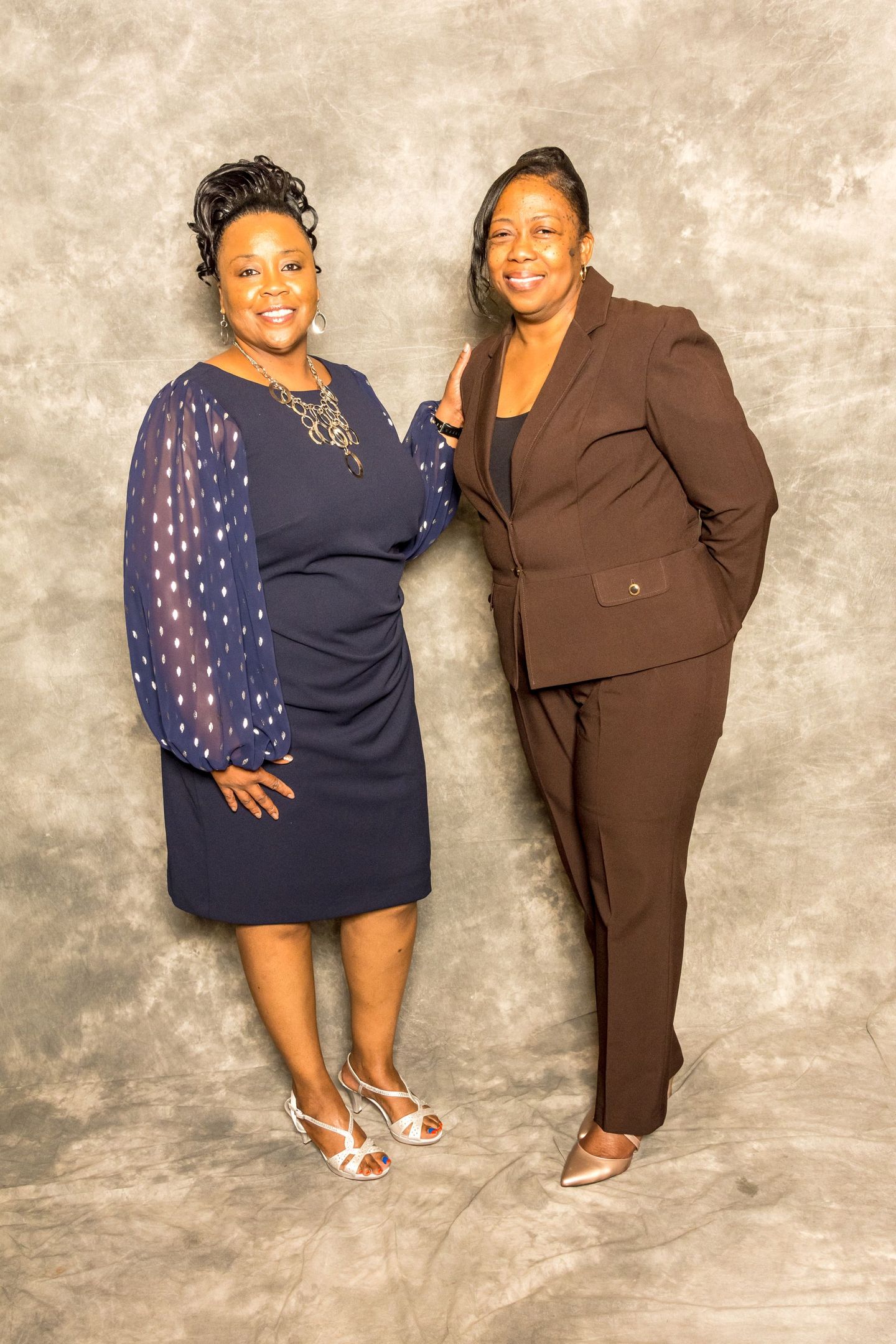 Two women posing together in formal outfits against a gray studio backdrop.