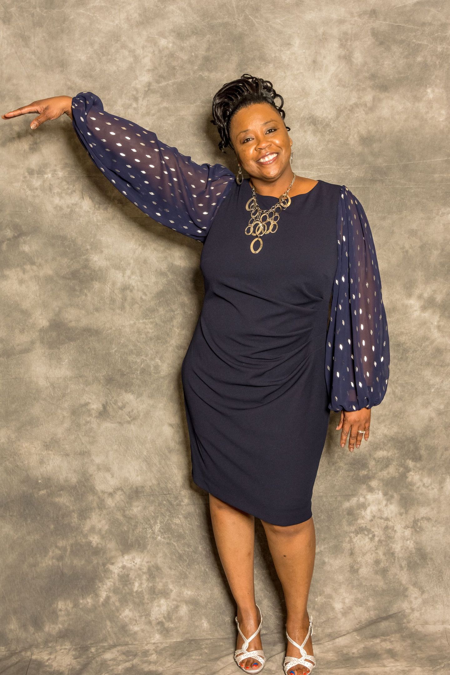 Woman in a navy dress posing with one arm raised against a gray backdrop.