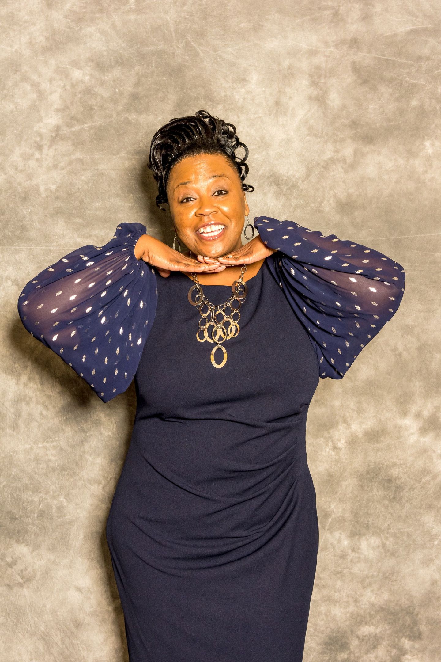 Woman in a navy dress with sheer polka-dot sleeves, posing against a neutral studio backdrop.
