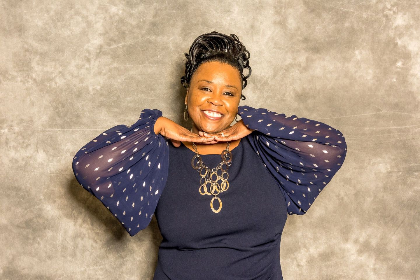Smiling woman posing with hands under chin against a gray backdrop, wearing a navy top with sheer dotted sleeves.