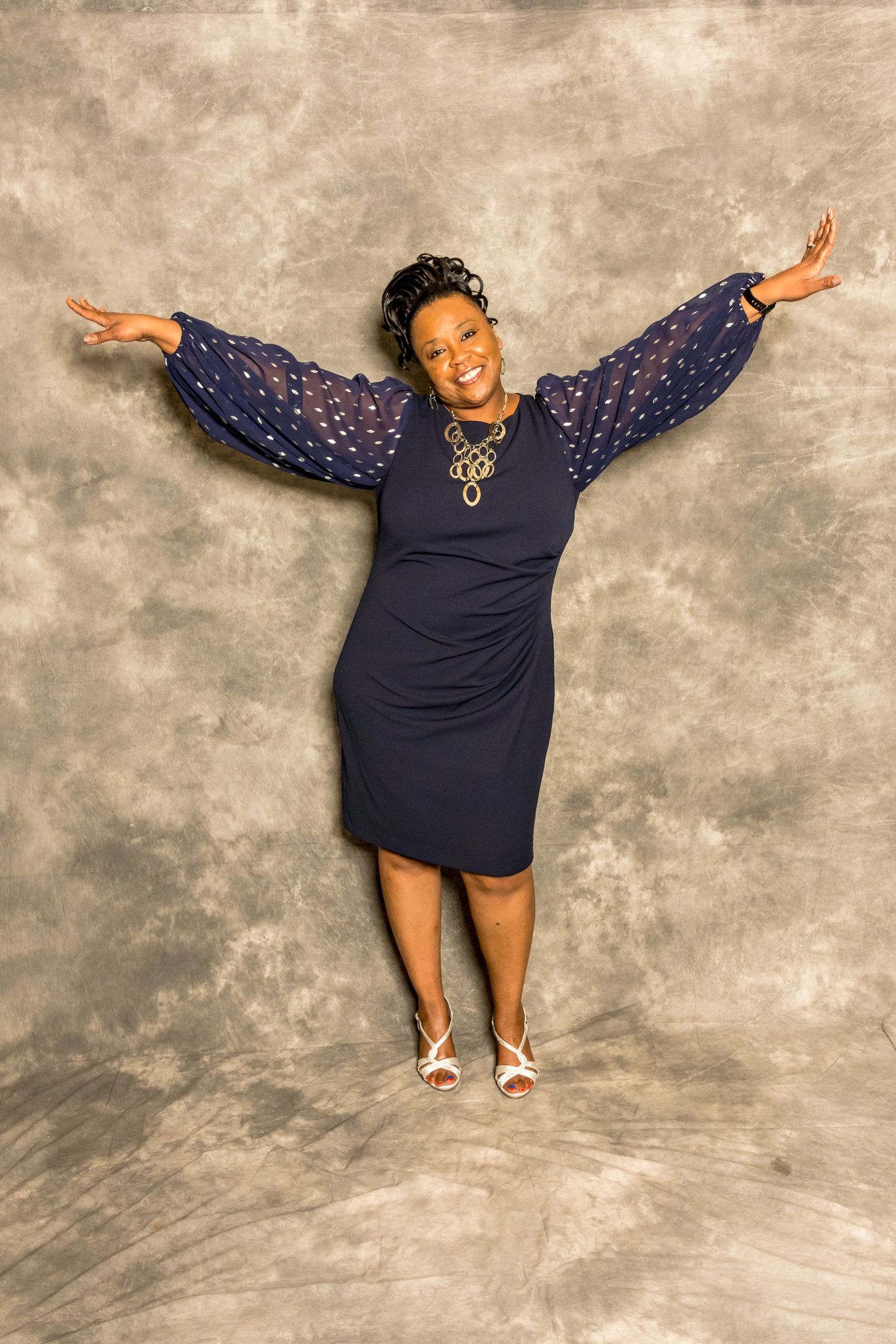 Person in a navy dress with sheer sleeves, smiling and holding arms out against a gray backdrop.