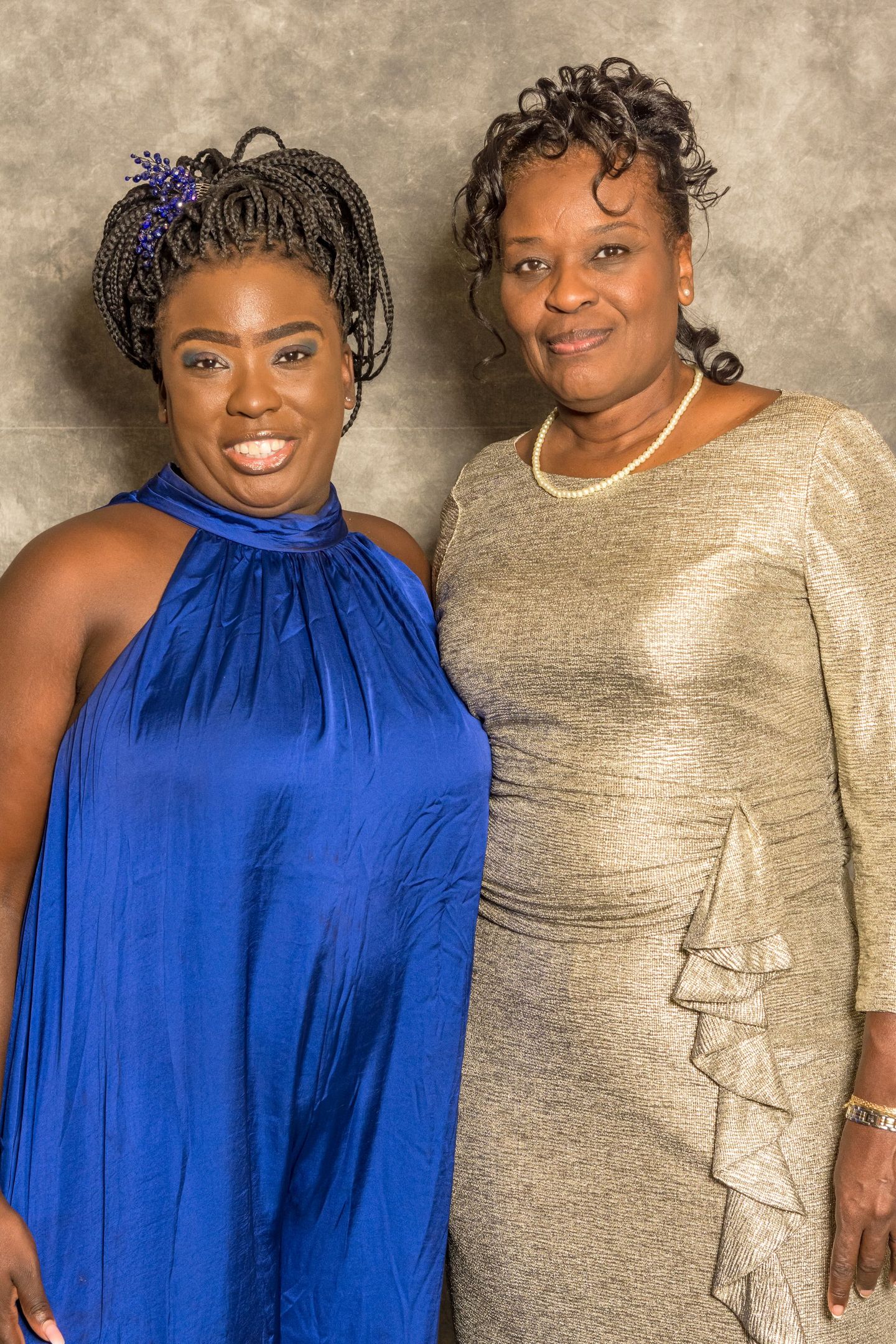 Two women posing together, one in a royal blue dress and one in a gold dress, smiling against a neutral backdrop.