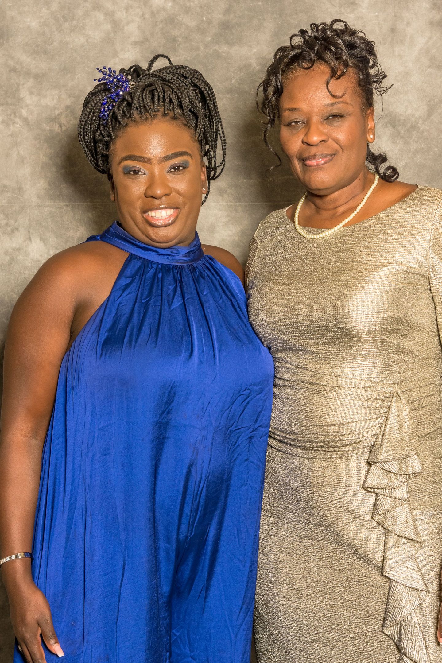 Two women posing together, one in a bright blue dress and one in a gold dress, smiling against a neutral backdrop