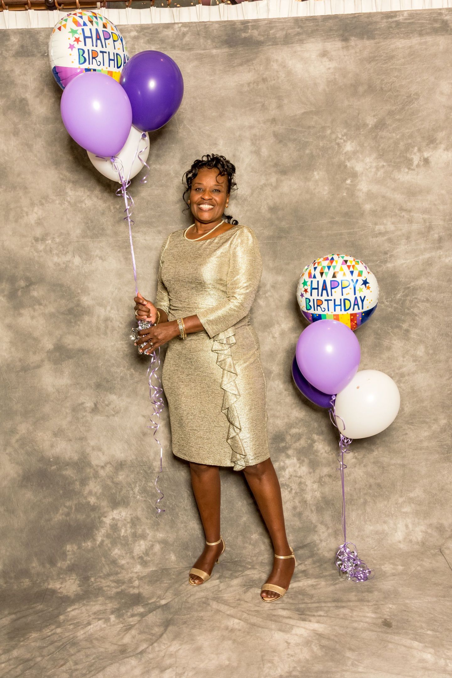 Woman in gold dress holding purple and white balloons against a gray backdrop