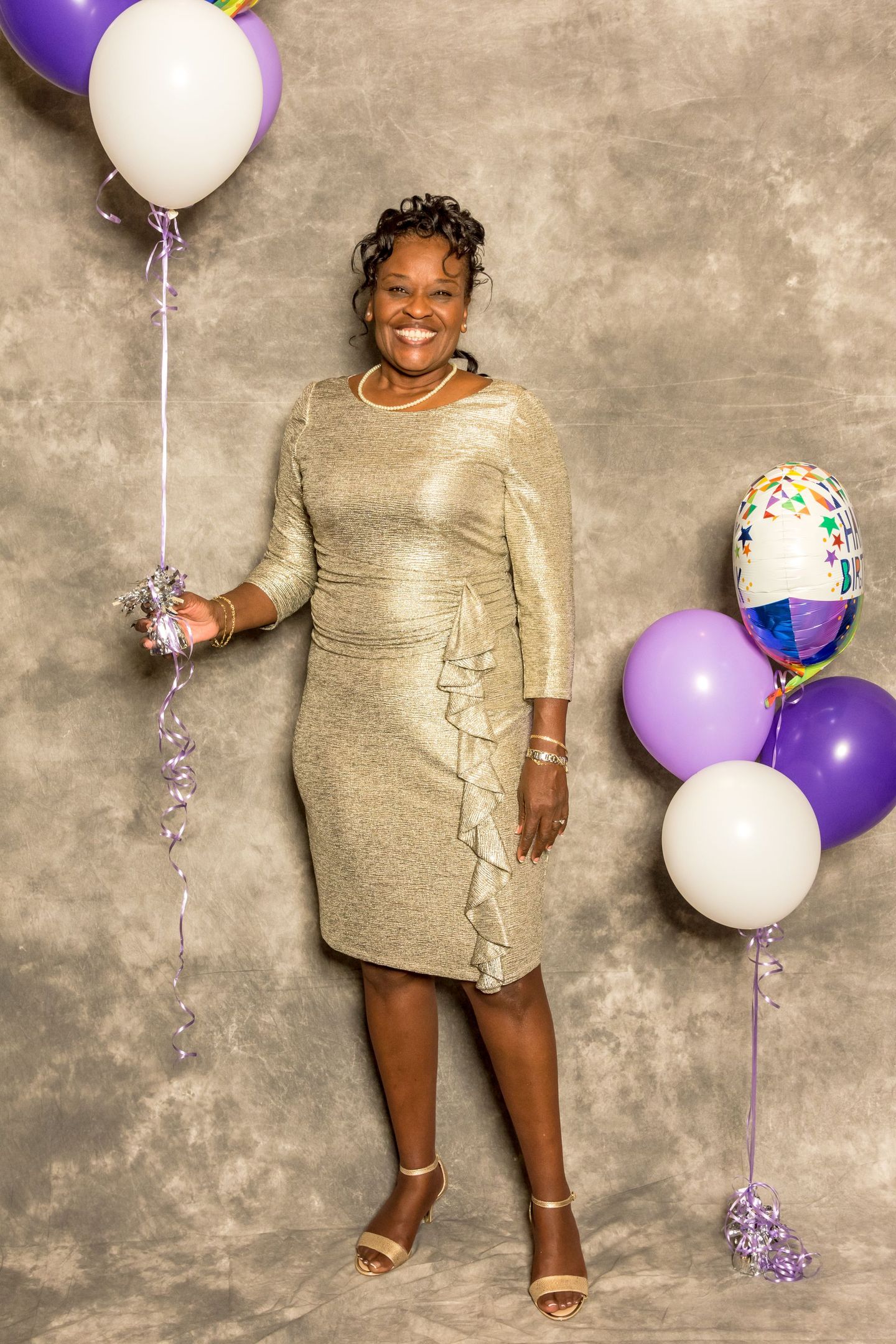 Person smiling in a gold dress, standing against a gray backdrop with purple and white balloons.