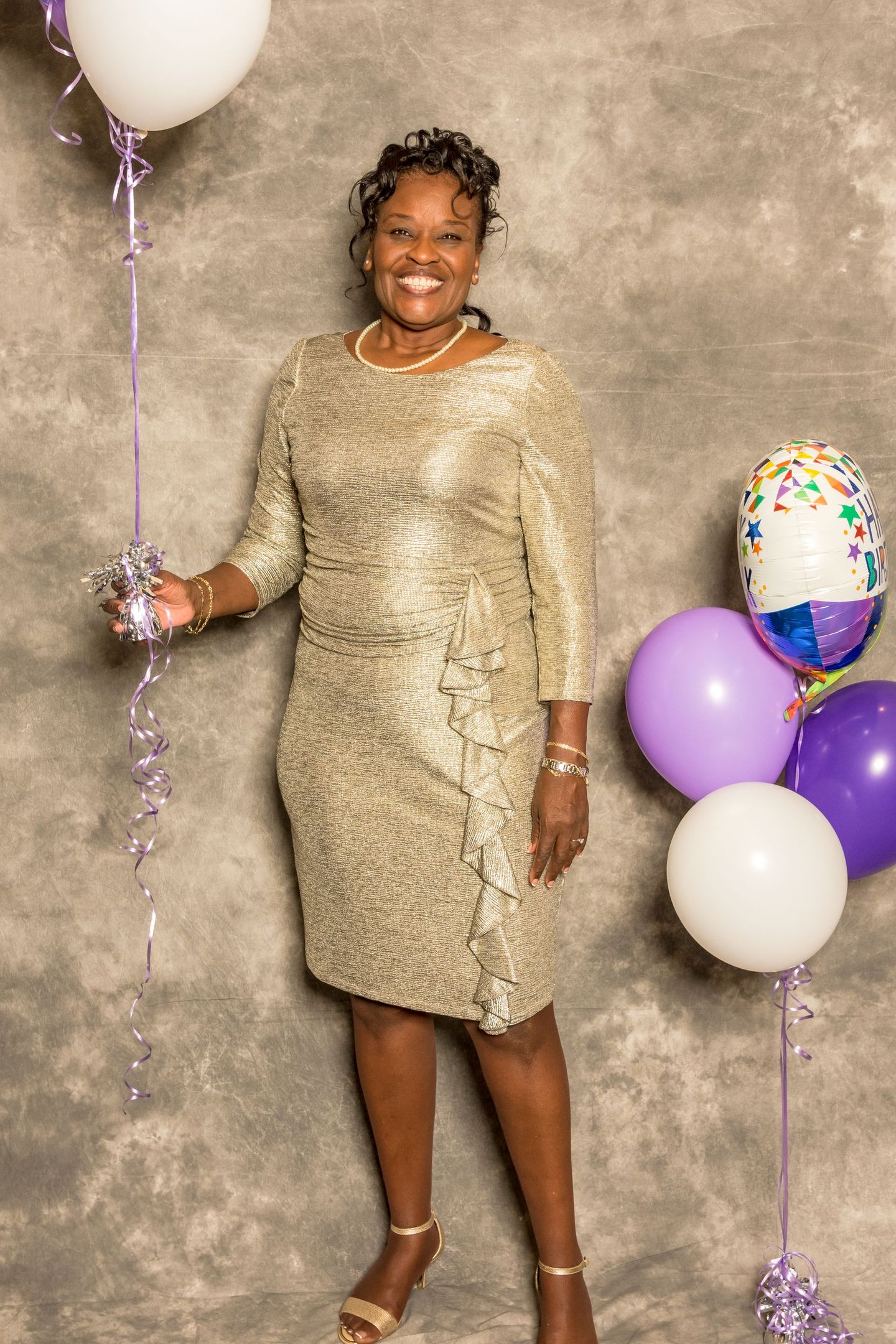 Woman in a gold dress standing by purple and white balloons against a gray backdrop