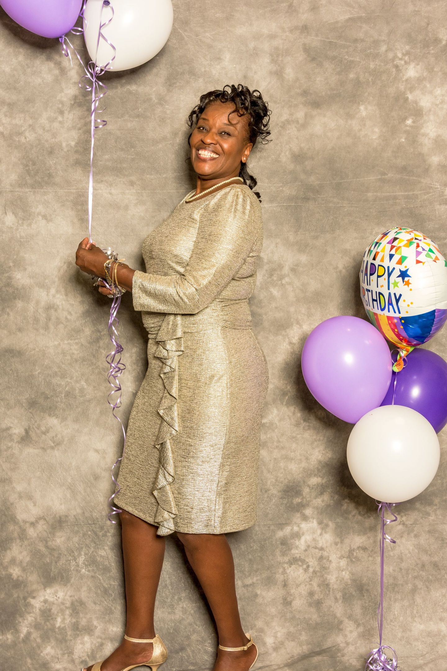 Woman in a gold dress smiling beside purple and white balloons against a textured backdrop.