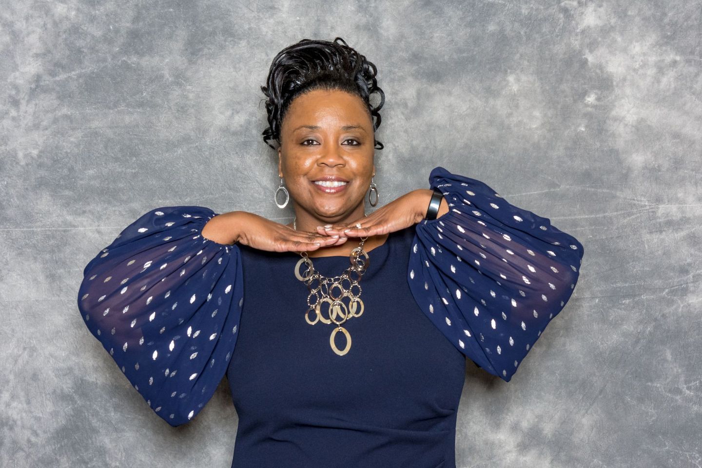 Smiling woman in a navy dress with sheer polka-dot sleeves, posing with hands under chin against a gray backdrop
