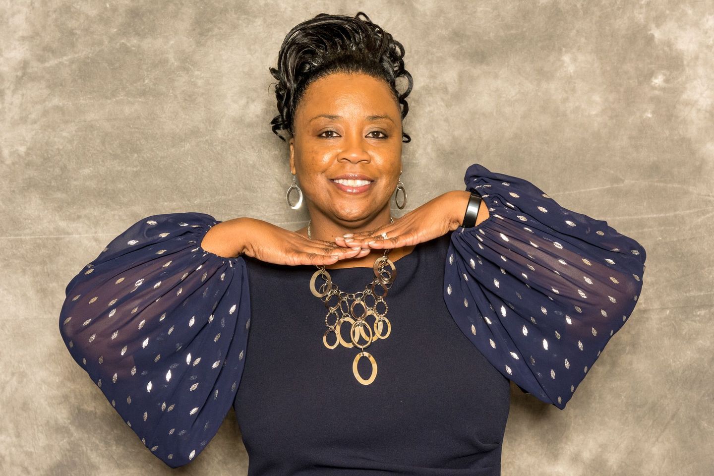 Woman posing against a gray backdrop in a navy top with sheer polka-dot sleeves and gold jewelry
