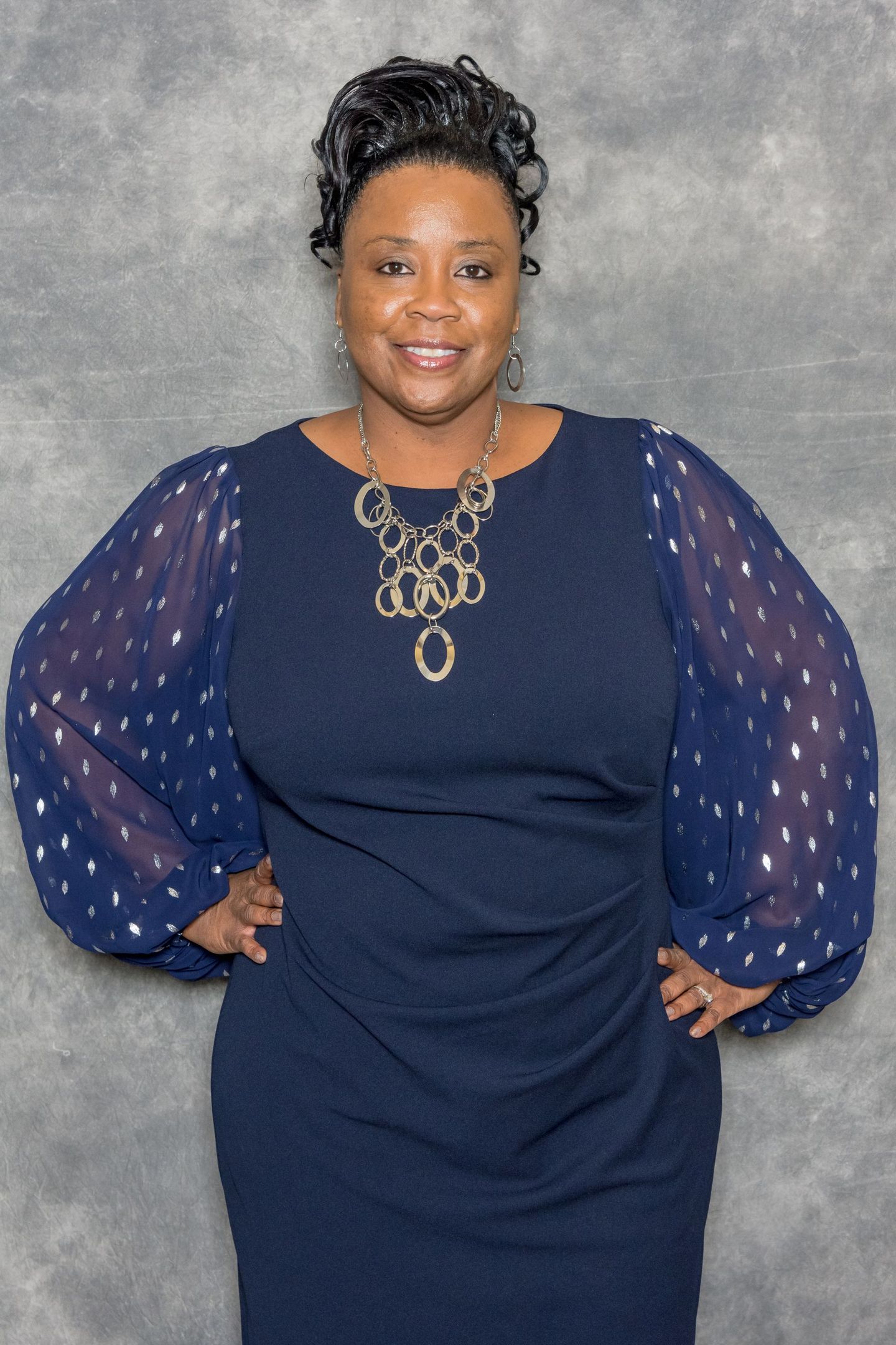 Woman in navy dress with sheer sleeves, posing against a gray backdrop and smiling.